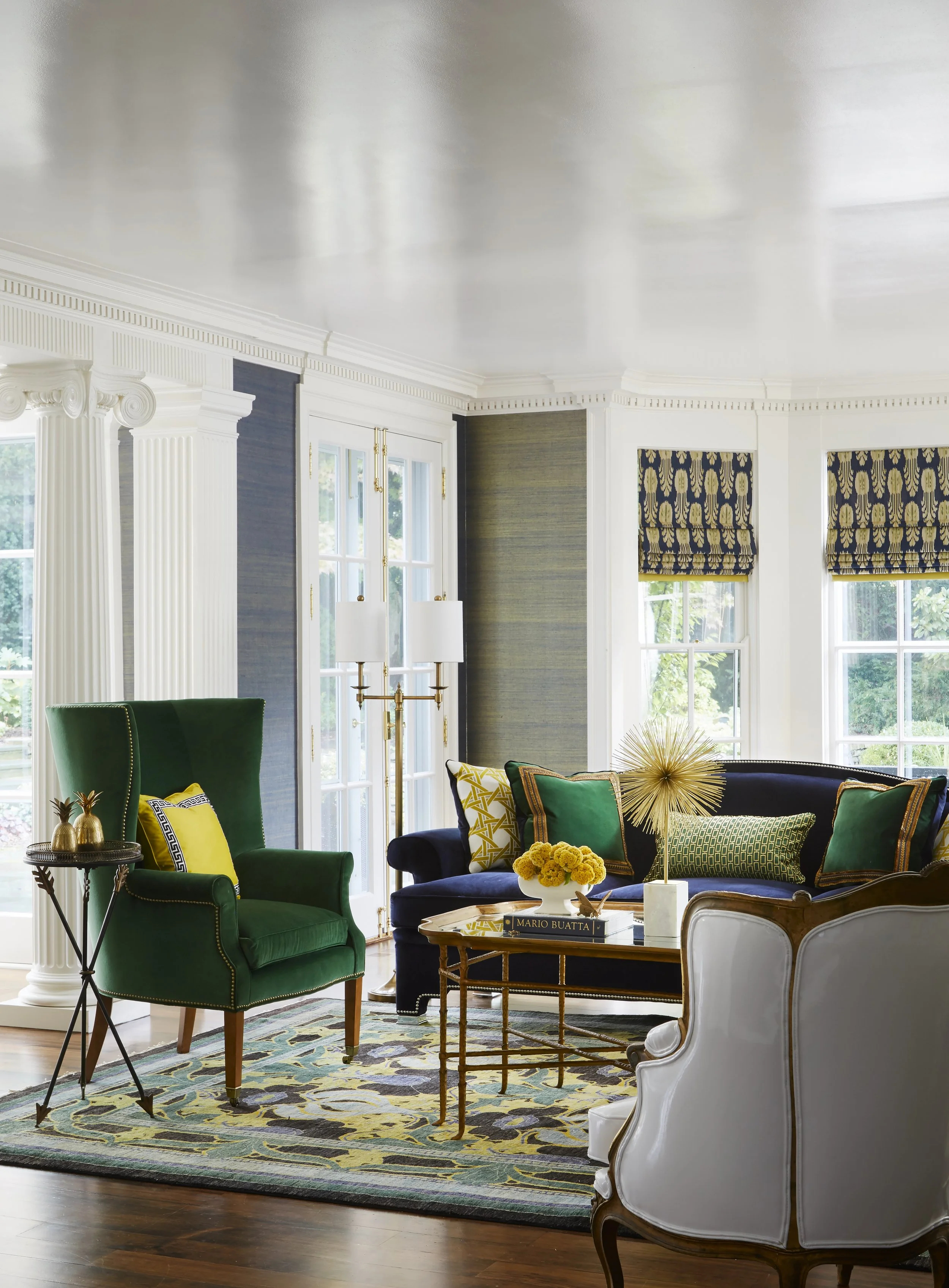 A bright living room with large windows, white and blue walls, green and dark blue upholstered furniture, decorative pillows, a gold starburst sculpture, a round coffee table with books and flowers, and a colorful patterned rug.