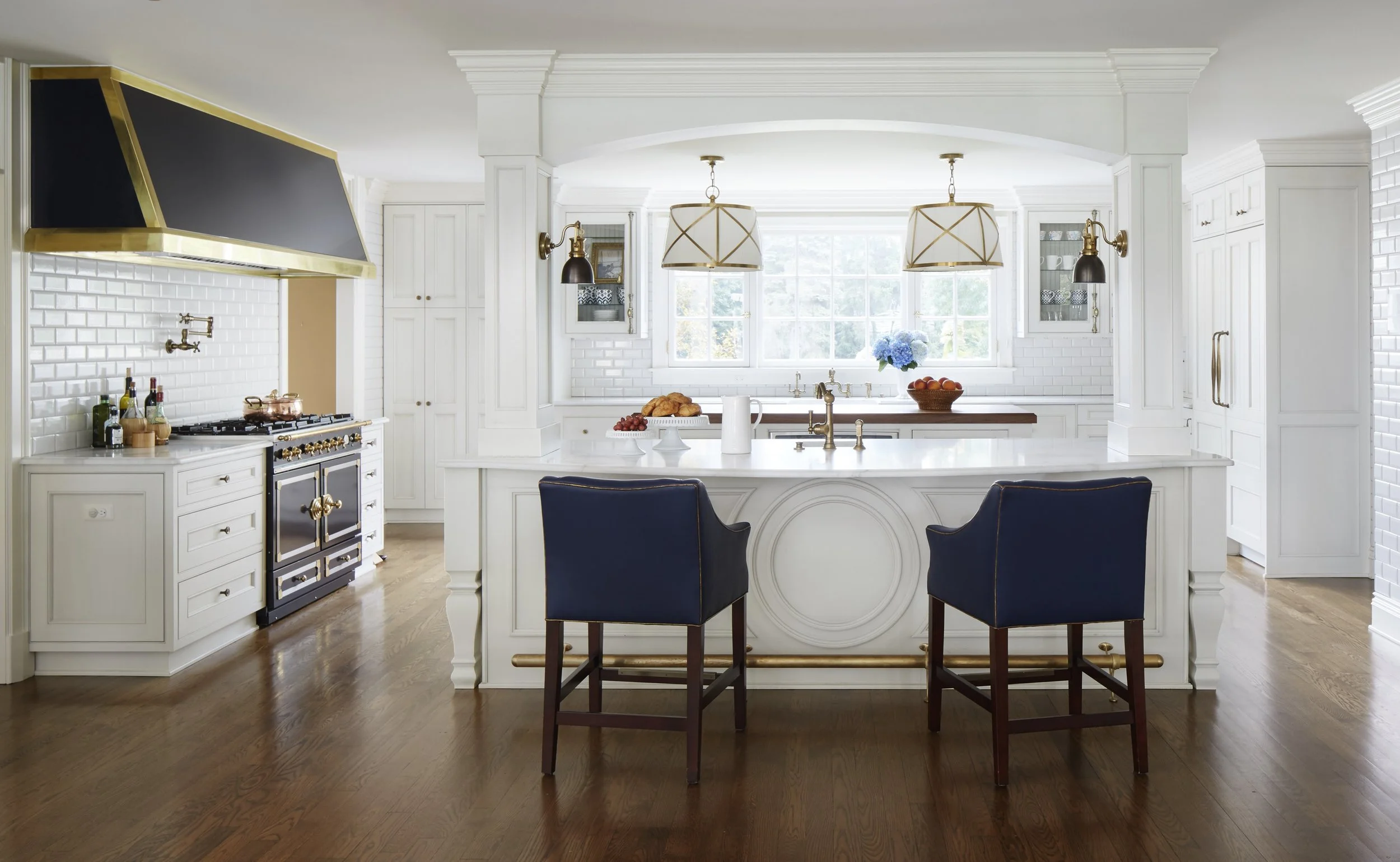 Bright, white kitchen with large window, white cabinets, and a central island with two navy blue chairs. There are two pendant lights hanging above the island, a flower arrangement, and a bowl of fruit on the window sill.