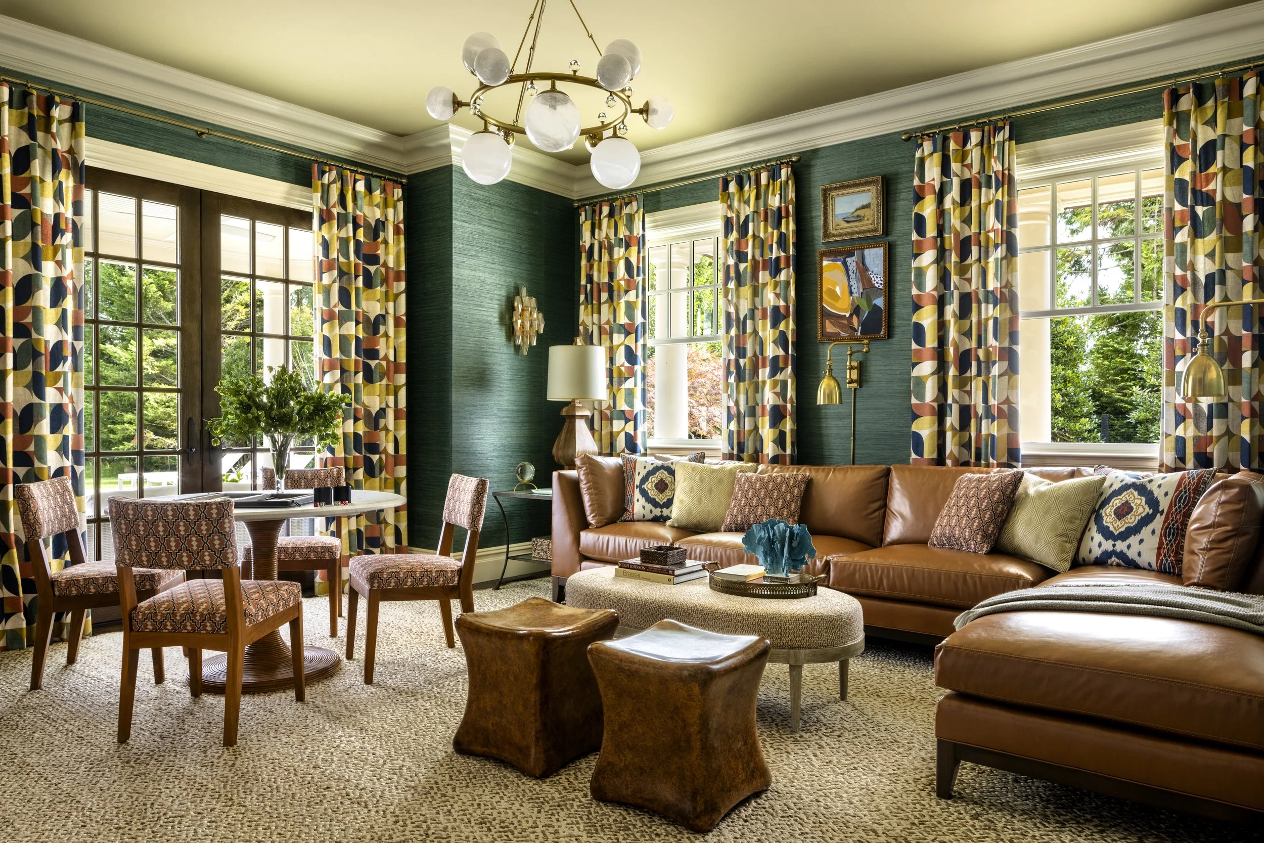 A bright living room with green textured walls, large windows with colorful geometric curtains, a brown leather sofa with patterned pillows, a round wooden dining table with chairs, a modern chandelier, and artwork on the walls.