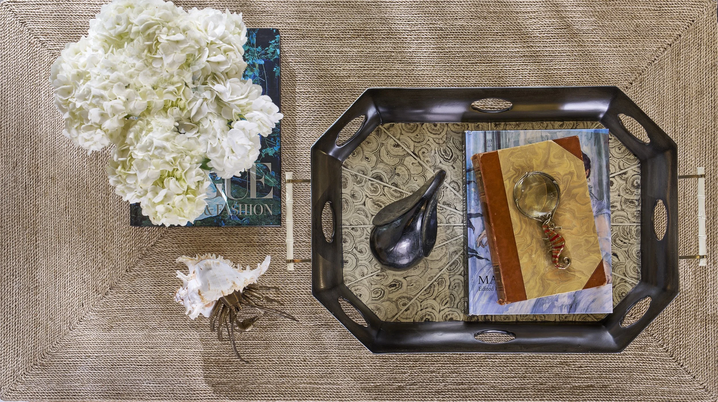 A black plastic tray table on a beige textured carpet with a bouquet of white hydrangeas, a magazine, a blue book, a brown book, a pair of glasses, a black shell, and a large conch shell.