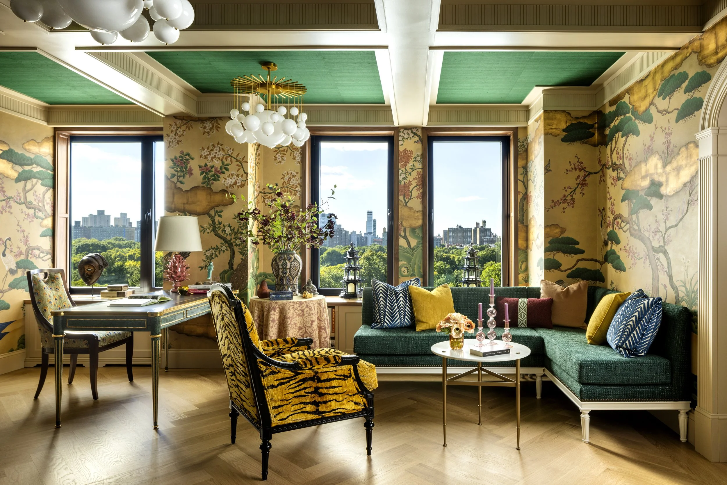Interior of a room with large windows showing a cityscape and greenery. The room features colorful furniture, including a green sofa with yellow and patterned pillows, a yellow and black animal print armchair, a wooden desk with a chair, various decorative items, and vases with plants. The ceiling has green panels and modern light fixtures.