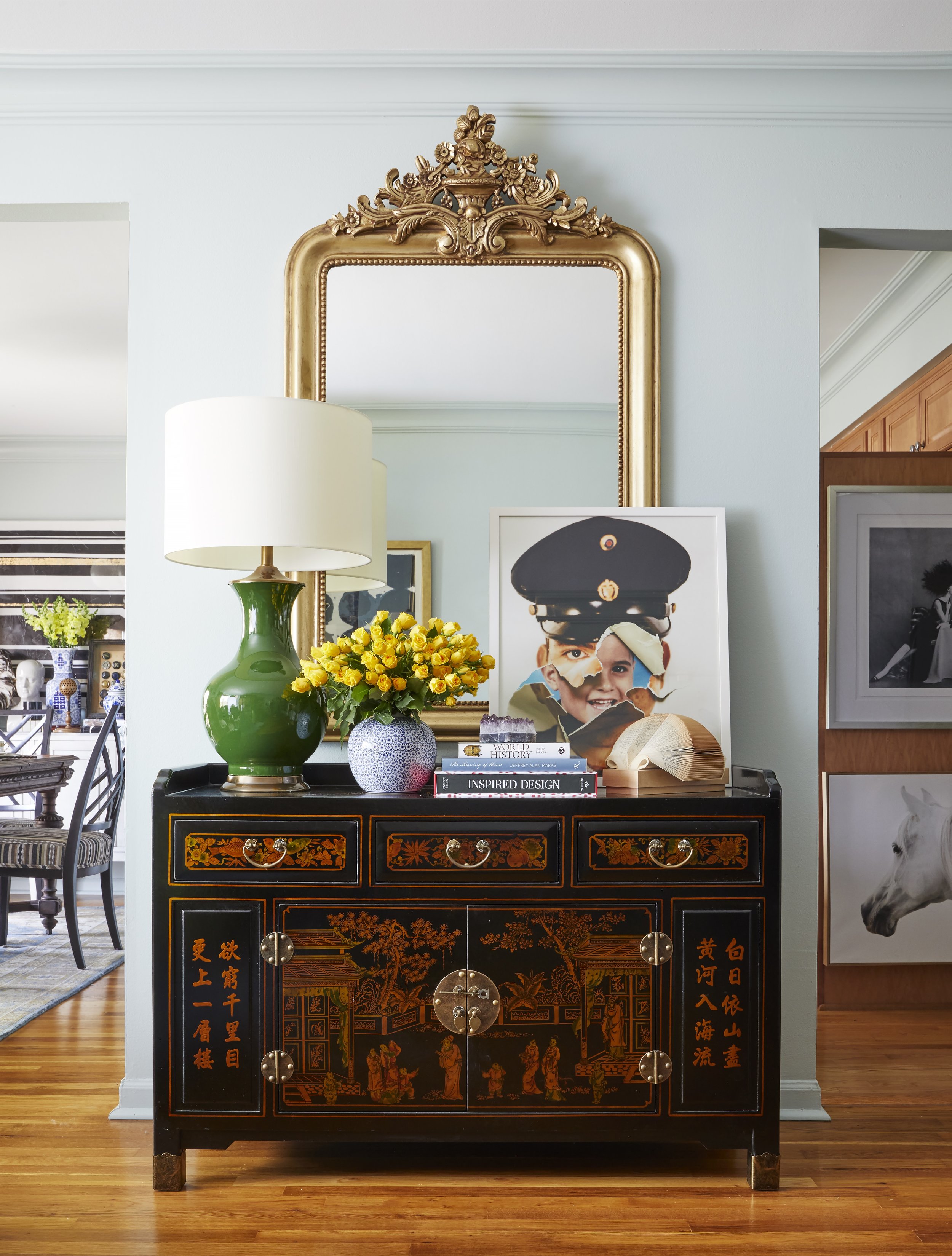 A black Chinese cabinet with colorful traditional artwork on the front, decorated with gold accents and hardware. It is topped with a large green vase holding a bouquet of yellow flowers, a small white and blue decorative bowl, a stack of books, and 