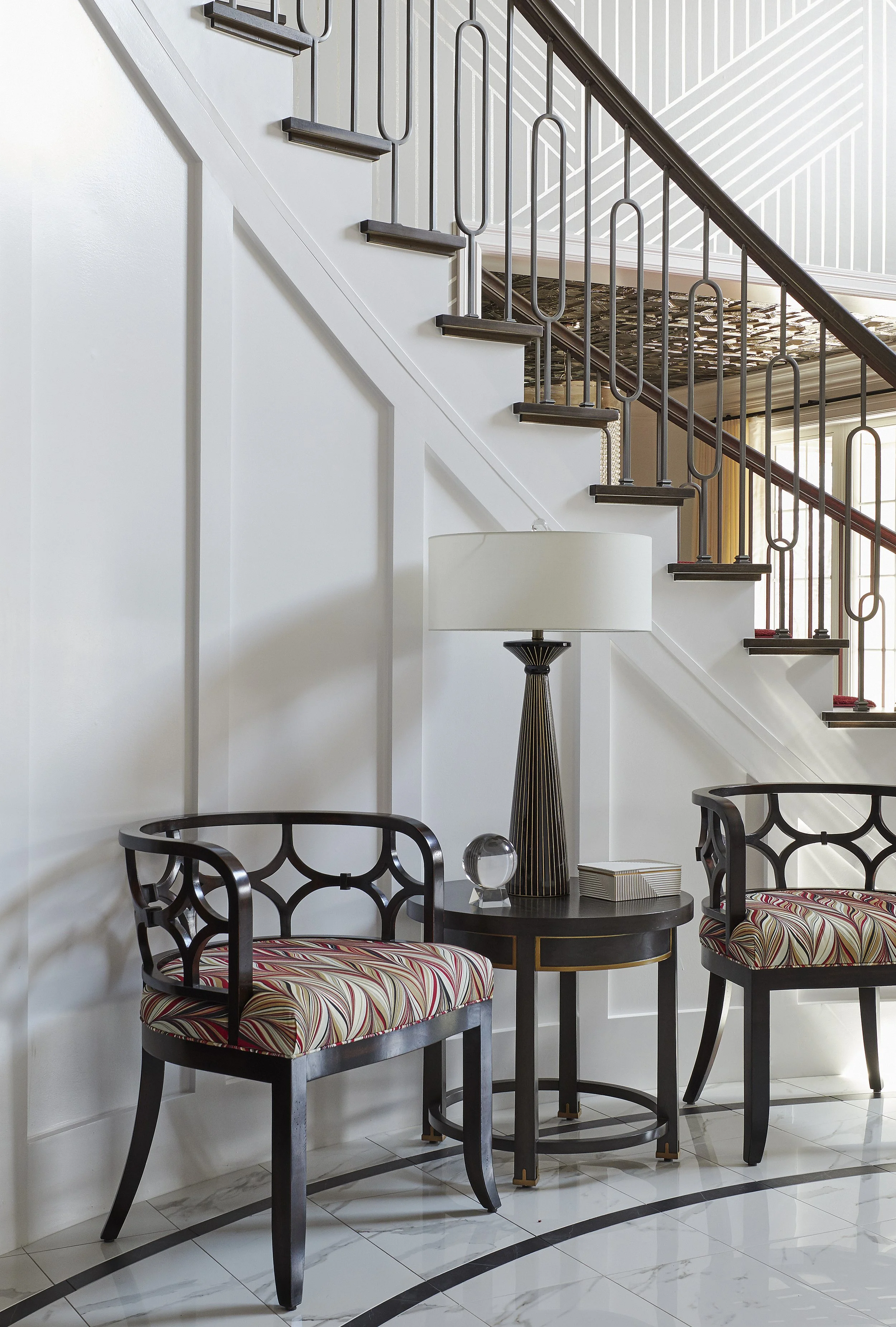 A modern, elegant interior with a staircase, a small round table, two black wooden chairs with patterned cushions, a tall striped lamp, and marble flooring with black accents.