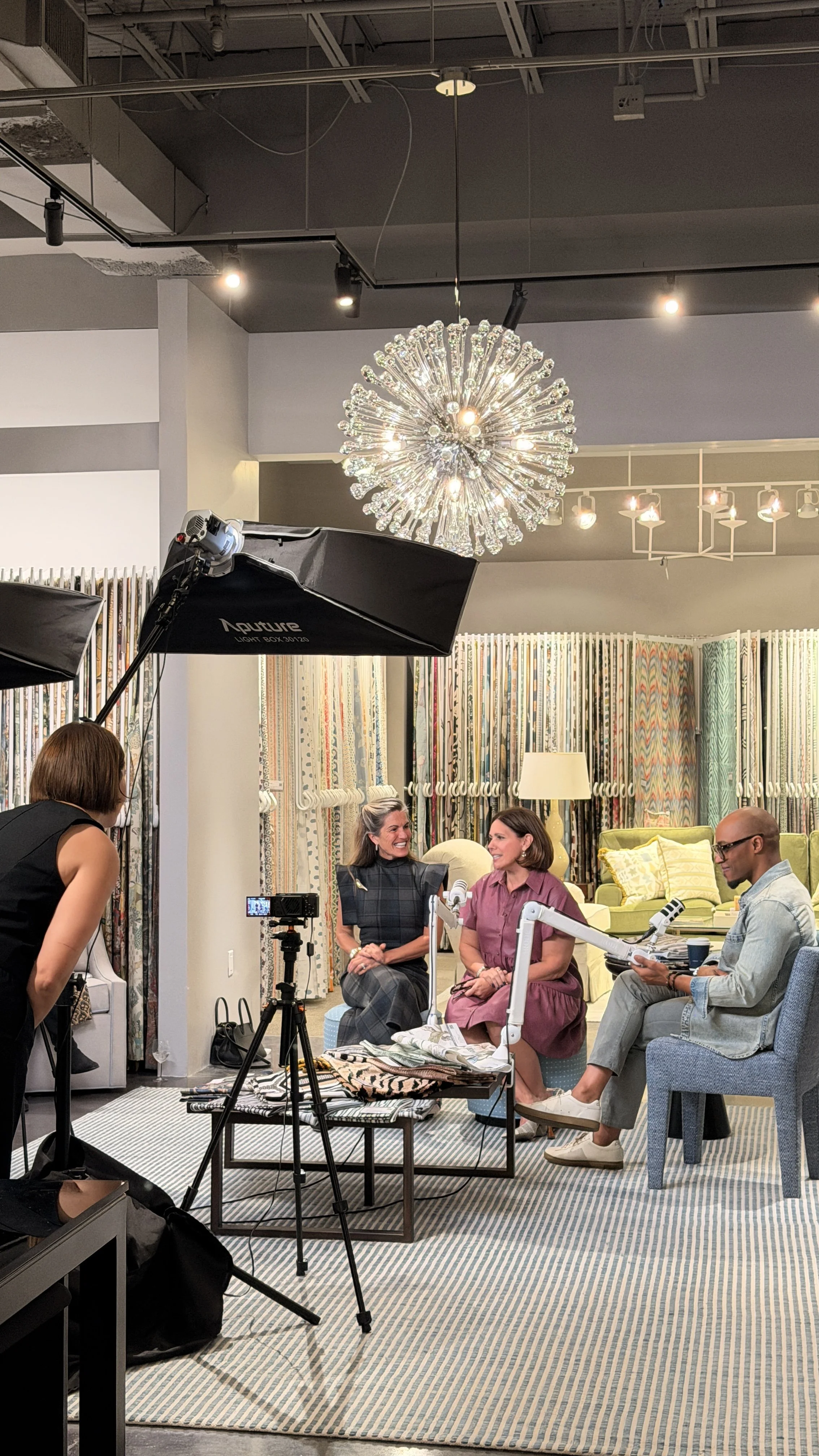 A group of four people participating in a recorded discussion or podcast in a stylish, well-lit room with colorful curtains, modern furniture, and decorative lighting fixtures. A woman in a black dress and a woman in a gray plaid dress are talking and smiling, while a man in a pink shirt and a man in a light blue shirt are listening.