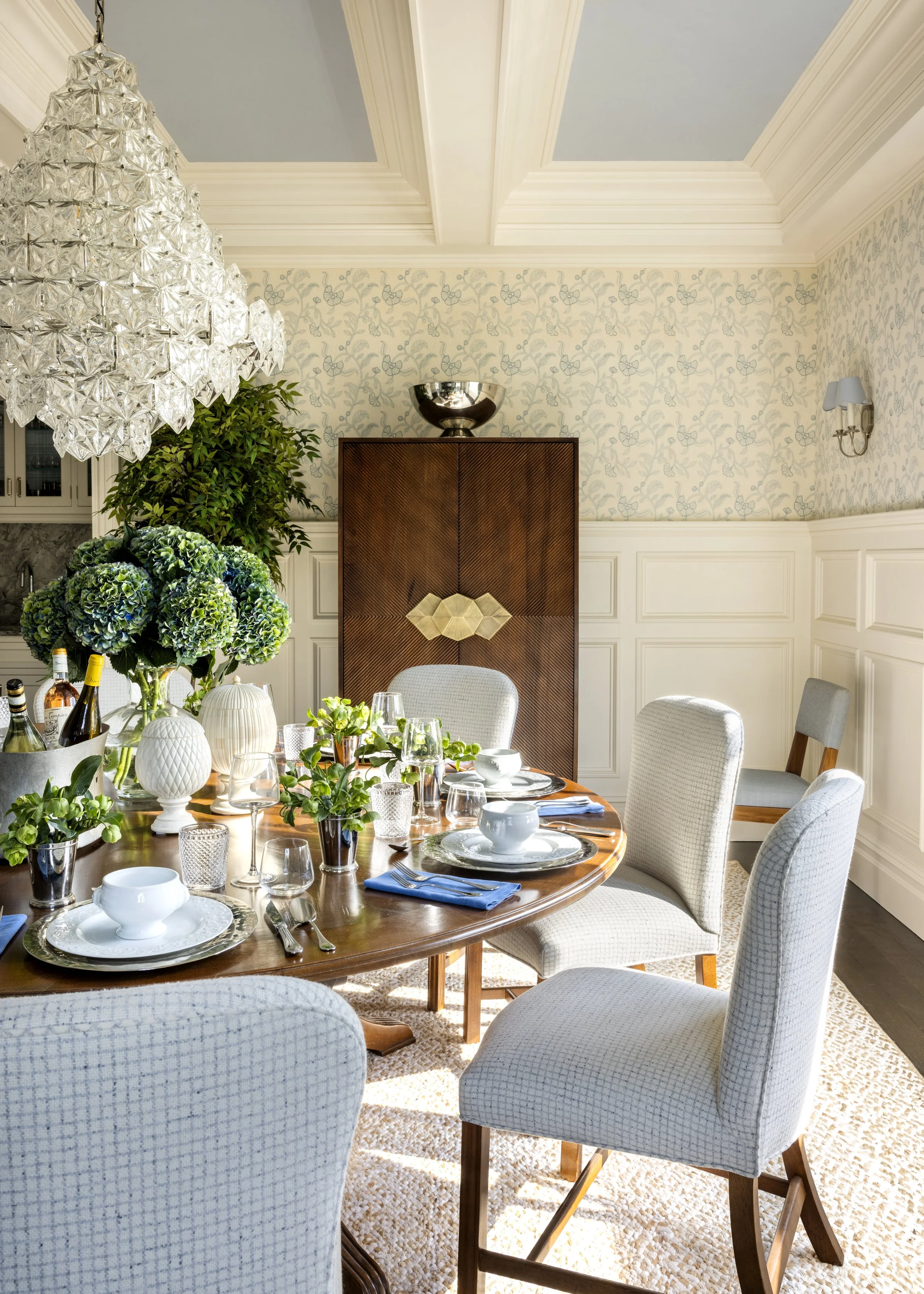 Elegant dining room with a round wooden table set with white dishes, glassware, and cutlery. The table is surrounded by upholstered chairs with light fabric. There are decorative vases and green plants on the table. A chandelier hangs from the ceilin