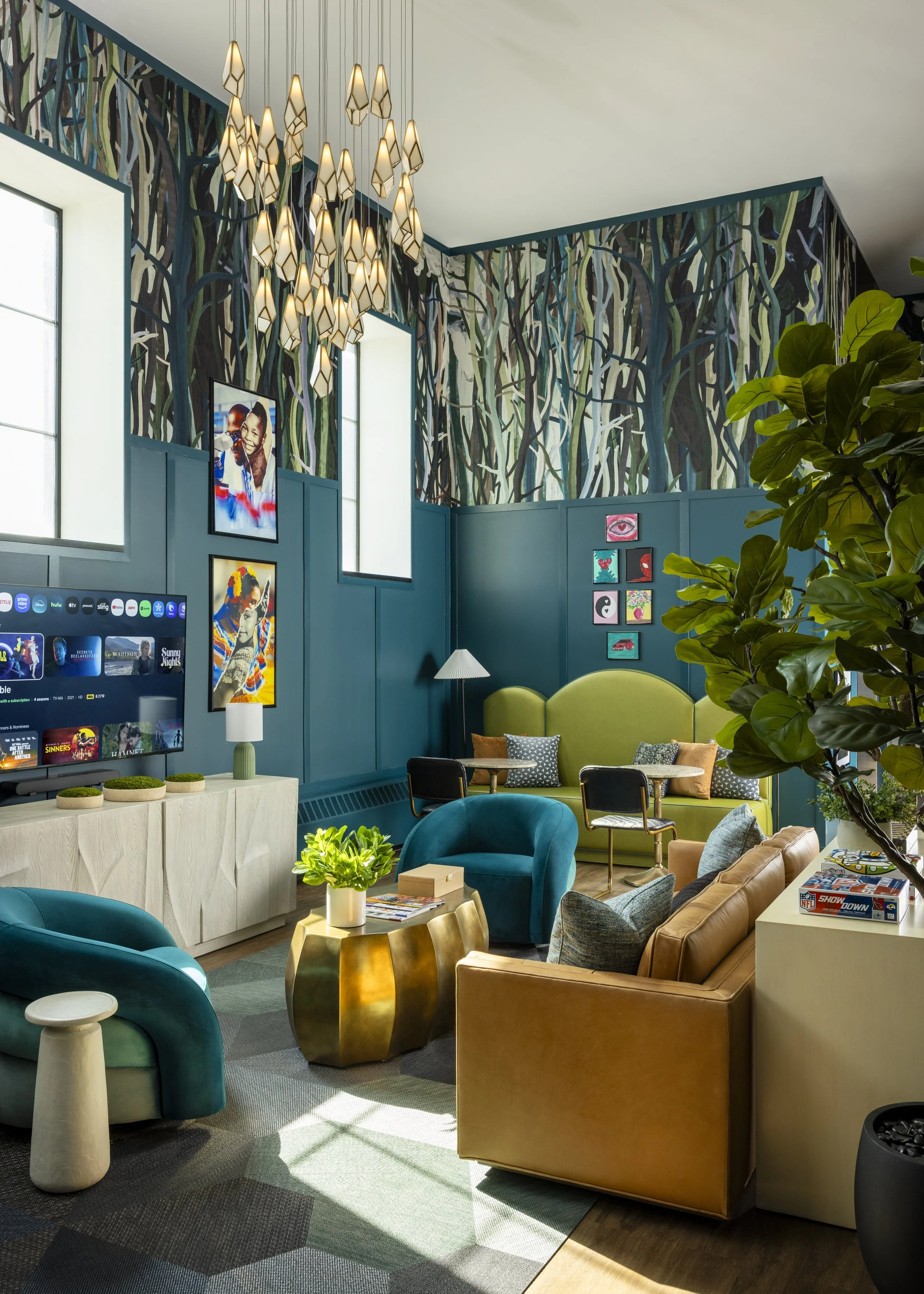 A colorful living room with a green, blue, and beige palette, featuring a large green sofa, a tan armchair, blue and teal chairs, and various small tables. The space includes vibrant wall art, a flat-screen TV, and a decorative chandelier with hangin