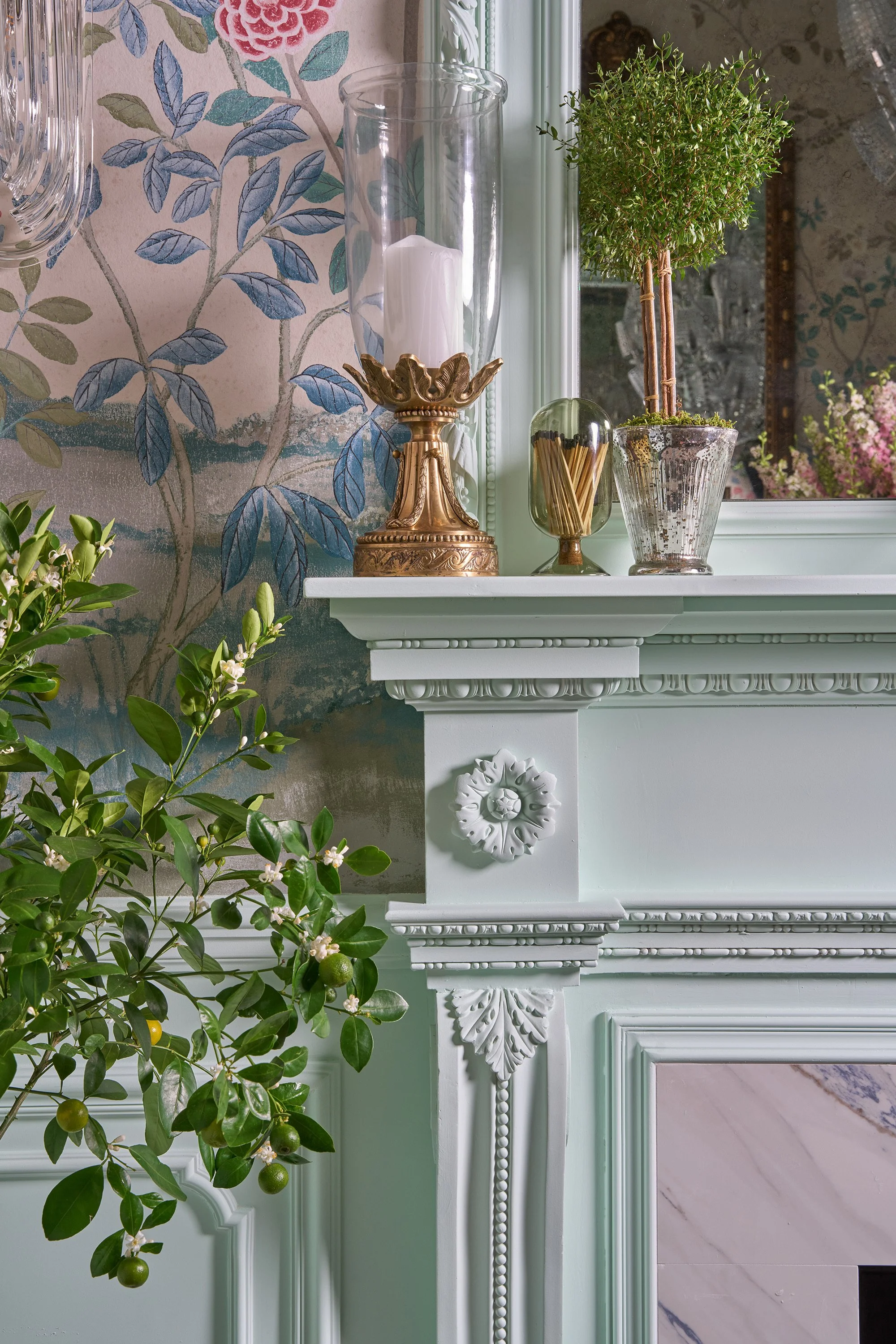 Decorative mantelpiece with a gold candle holder, a glass hurricane with a white candle, a small potted tree, and a jar of toothpicks. A part of a floral wallpaper and a mirror reflecting pink flowers are visible in the background, along with some gr