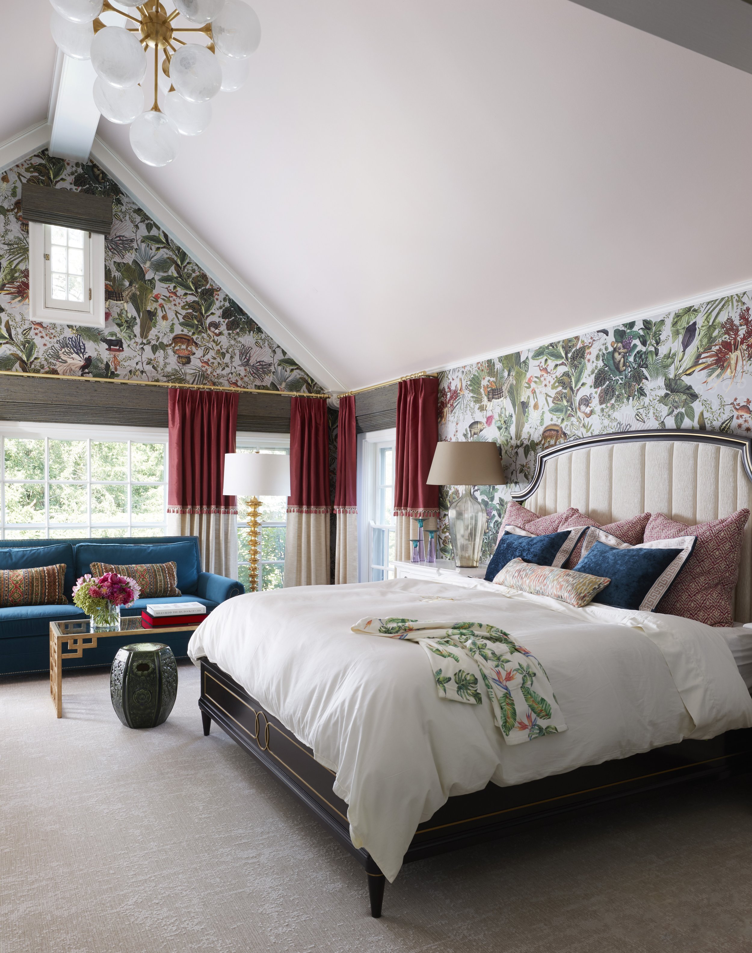A bedroom featuring a large bed with a beige upholstered headboard, multiple patterned pillows, and a white duvet, situated in front of a window with red curtains. A black lantern-style side table and a clear glass lamp with a beige shade are beside 