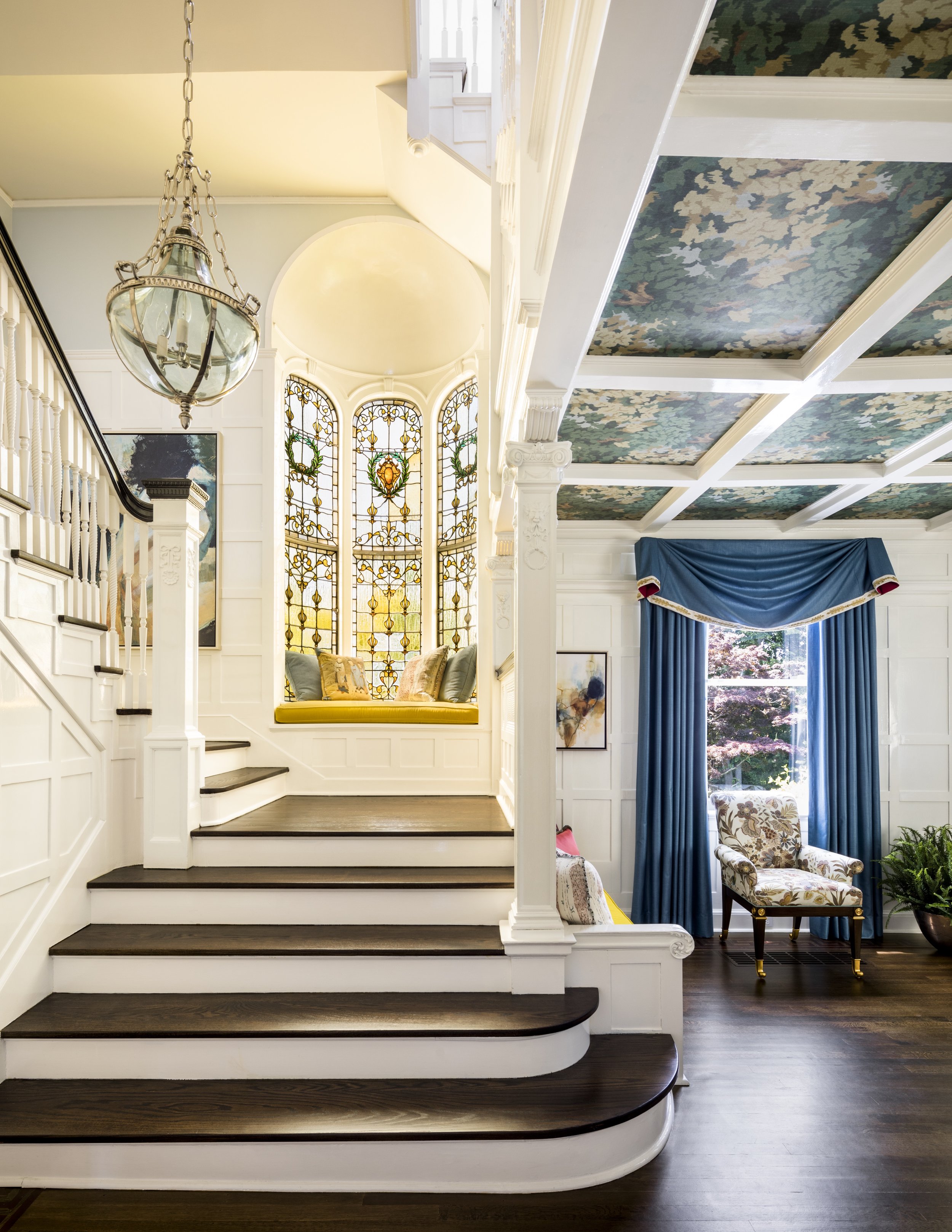 Interior view of a house featuring a staircase with dark wooden steps, a decorative stained-glass window with warm sunlight, and a cozy window seat with cushions. To the right, there's a sitting area with a floral-patterned armchair and deep blue cur