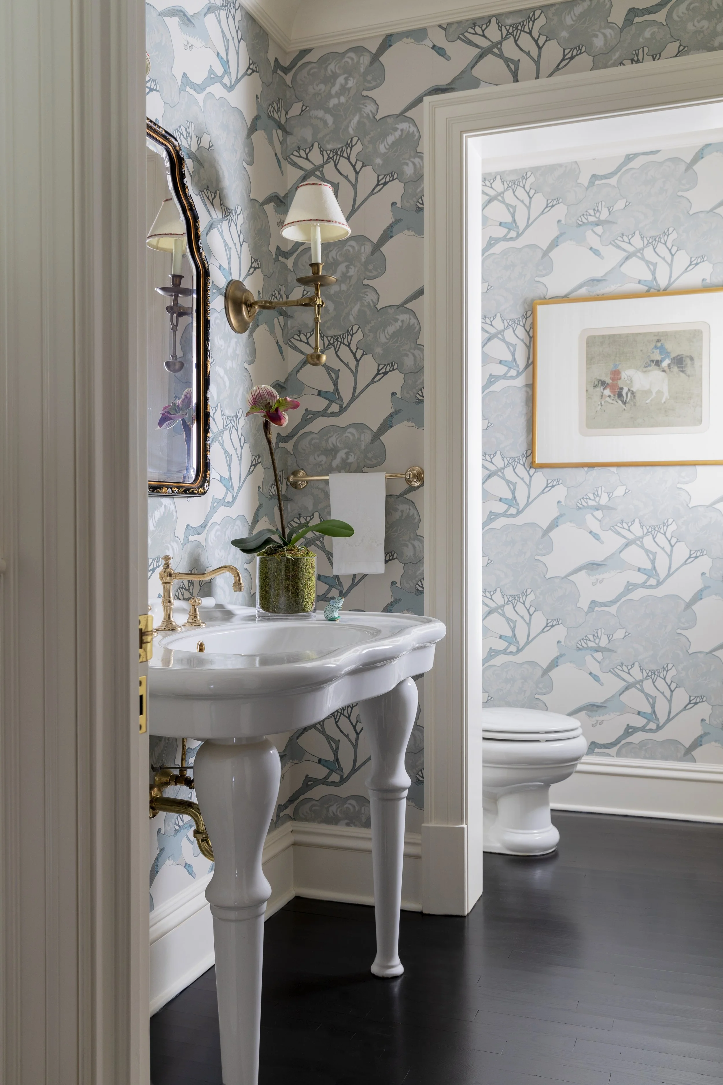 A vintage-style bathroom with a white pedestal sink, a large framed mirror, and a wall-mounted brass light fixture with two lampshades. The wall has wallpaper featuring a light blue and gray cloud and tree pattern. There is a framed picture of horses