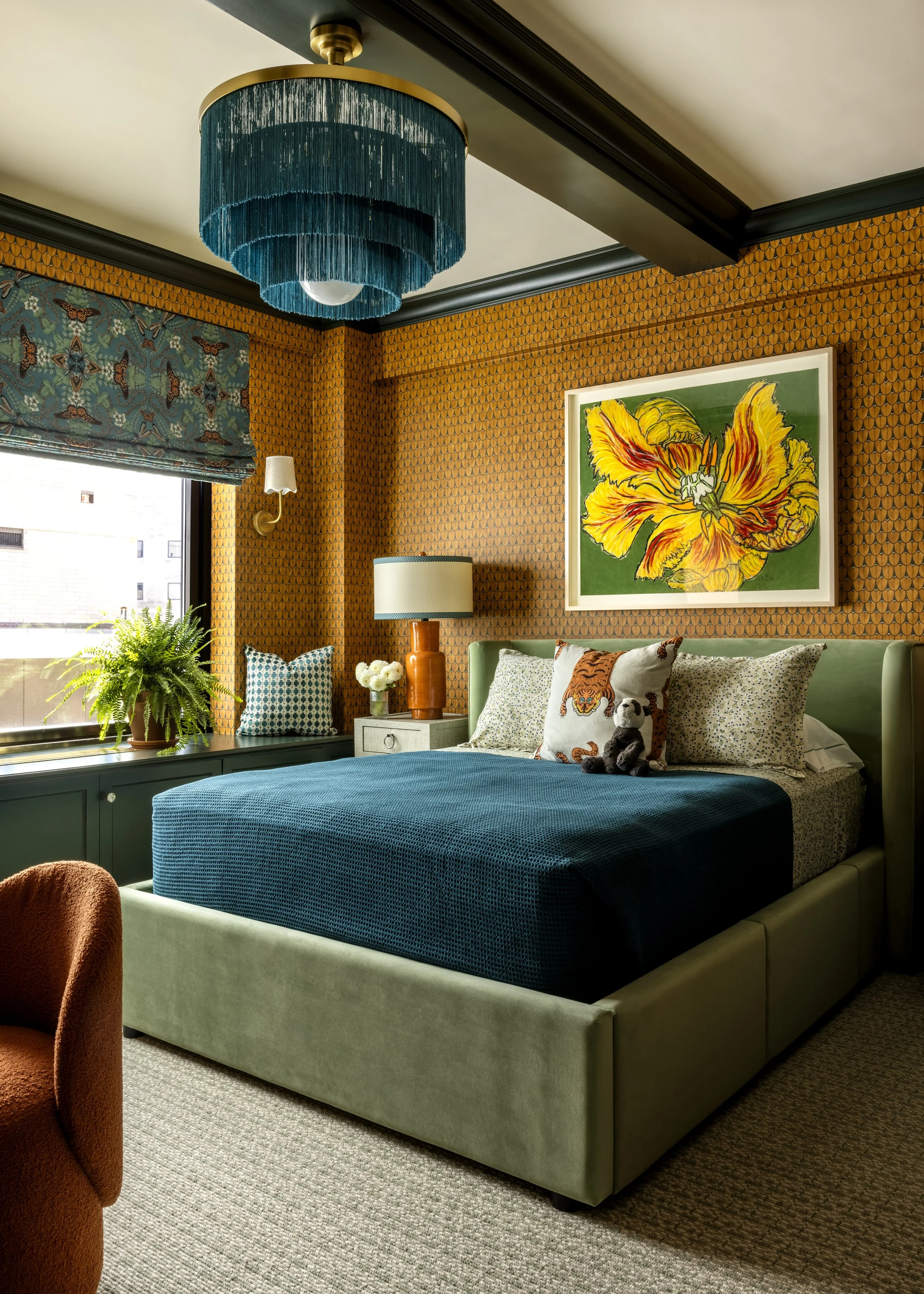 A bedroom with a green upholstered bed, a blue textured bedspread, patterned pillows, and a stuffed animal. Above the bed, a large framed flower artwork hangs on the orange patterned wall. There are windows with a teal and gold chandelier hanging from the ceiling, and a table lamp on a white nightstand. A plant sits by the window, and part of an orange upholstered chair is visible in the foreground.