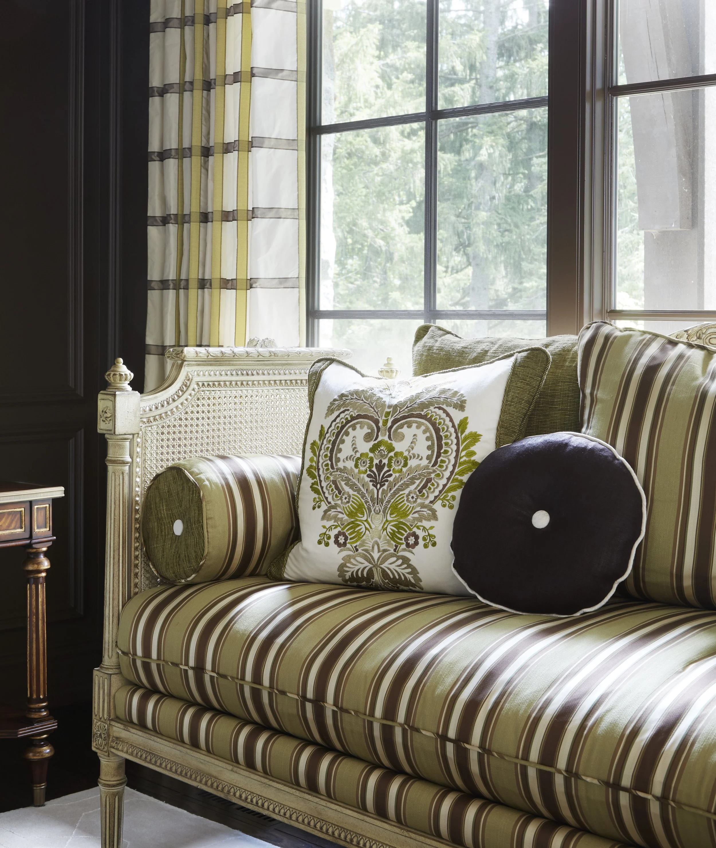 Interior of a room featuring a vintage cream-colored wooden sofa with striped upholstery in green, brown, and beige. The sofa is decorated with patterned and plain cushions, including a white cushion with a green and brown floral design and a black r