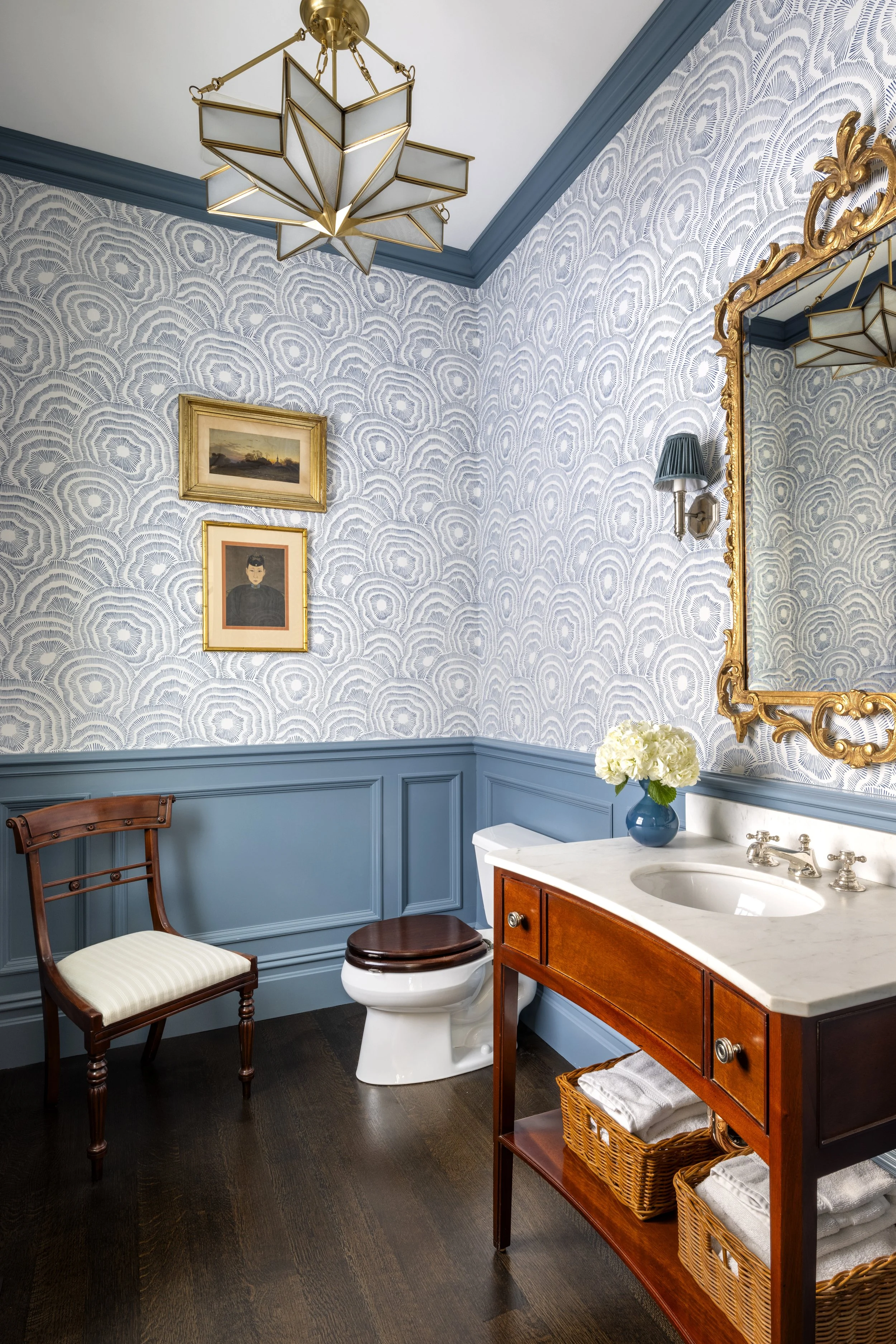 A vintage-style bathroom with blue patterned wallpaper, a gold-framed mirror, a wall-mounted lamp with a blue shade, a small sink with a wooden vanity, a chair, a toilet, a vase with white flowers, and framed artwork on the wall.