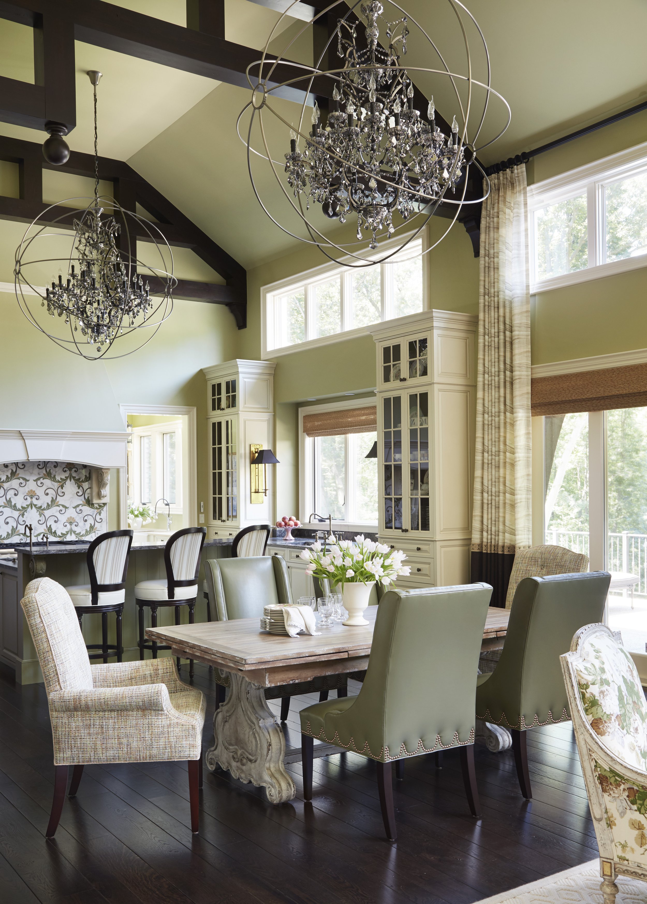A spacious dining area with a rustic wooden table surrounded by a mix of upholstered and wooden chairs. A large vase with white flowers is on the table, and a fireplace with a decorative tile backsplash is in the background. Large windows with natura