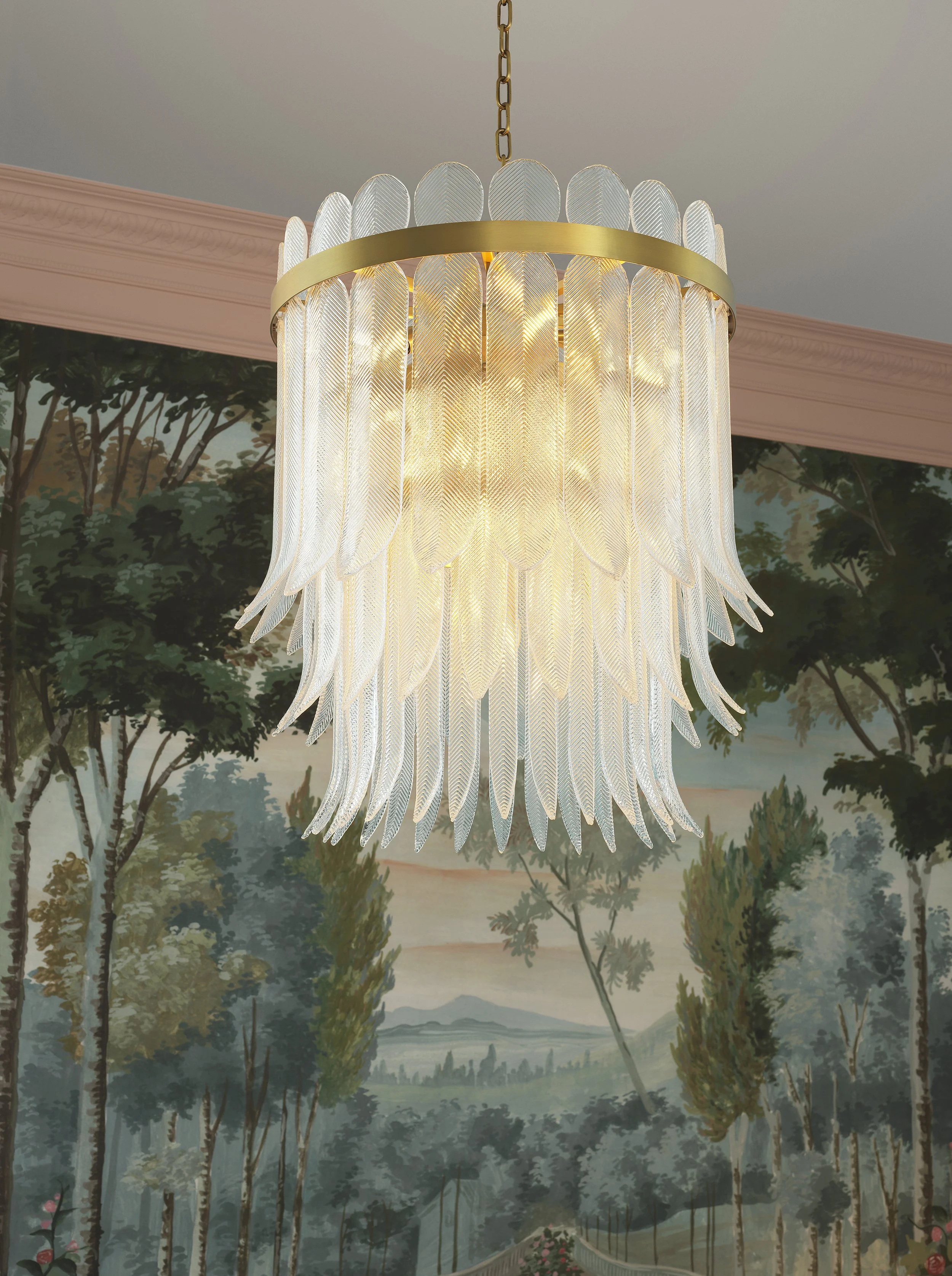 A large, ornate chandelier with many glass feathers hangs from a gold chain, illuminating a room with a landscape mural of trees and a pathway.