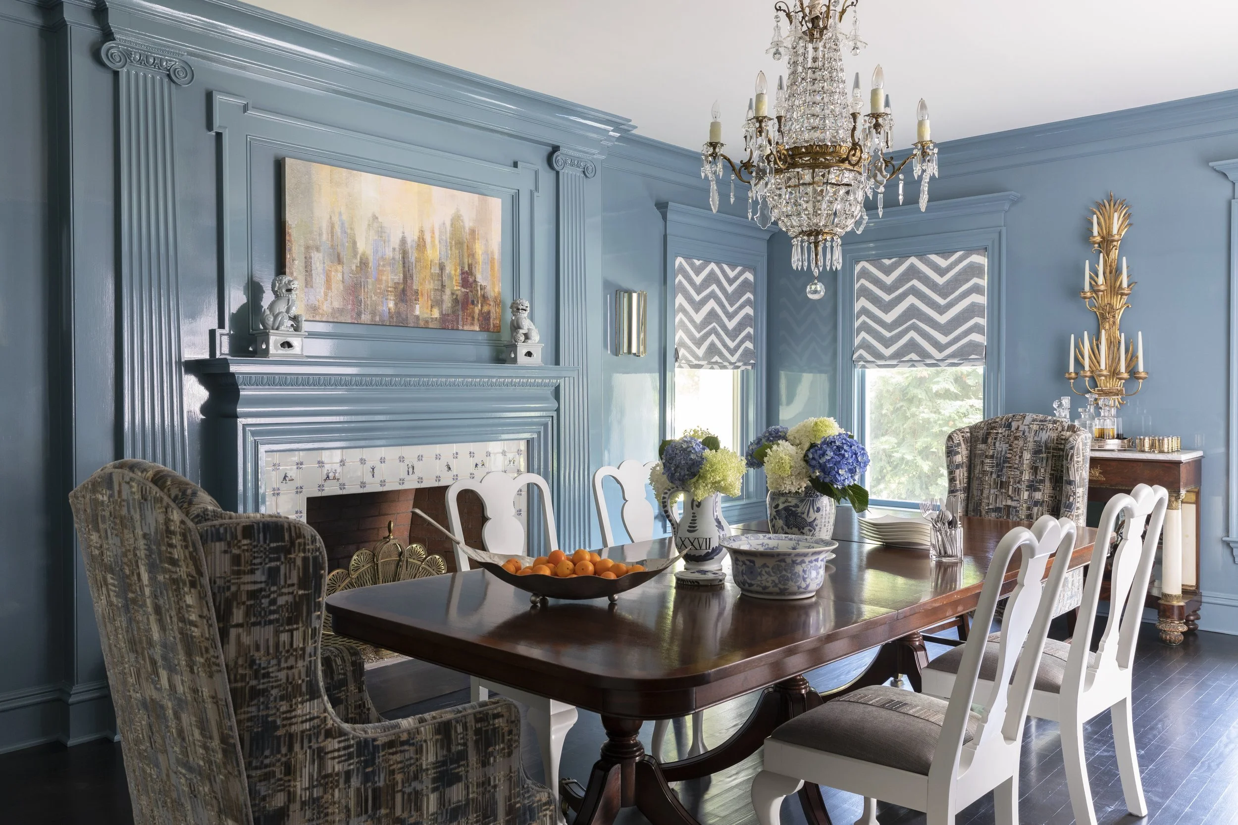 A dining room with blue walls, a wooden dining table, white and upholstered chairs, a chandelier, and a fireplace with a colorful cityscape painting above it. The table is set with bowls of fruit and decorative vases.