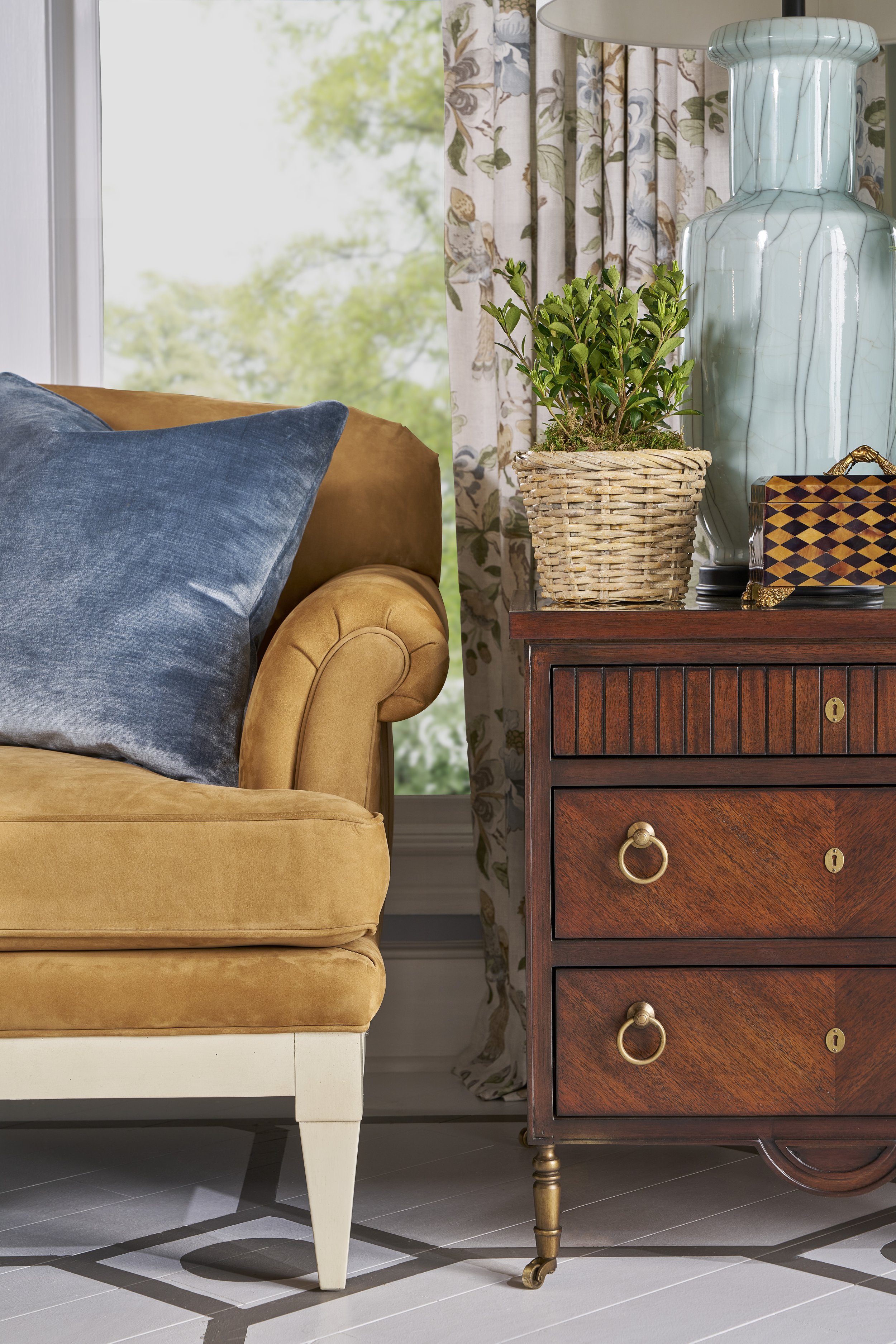 Close-up of a mustard-colored upholstered sofa with a blue velvet pillow. A wooden side table with drawers and gold ring pulls is beside it, decorated with a wicker basket with a green plant, a blue vase with a crackled glaze, and a decorative box. A