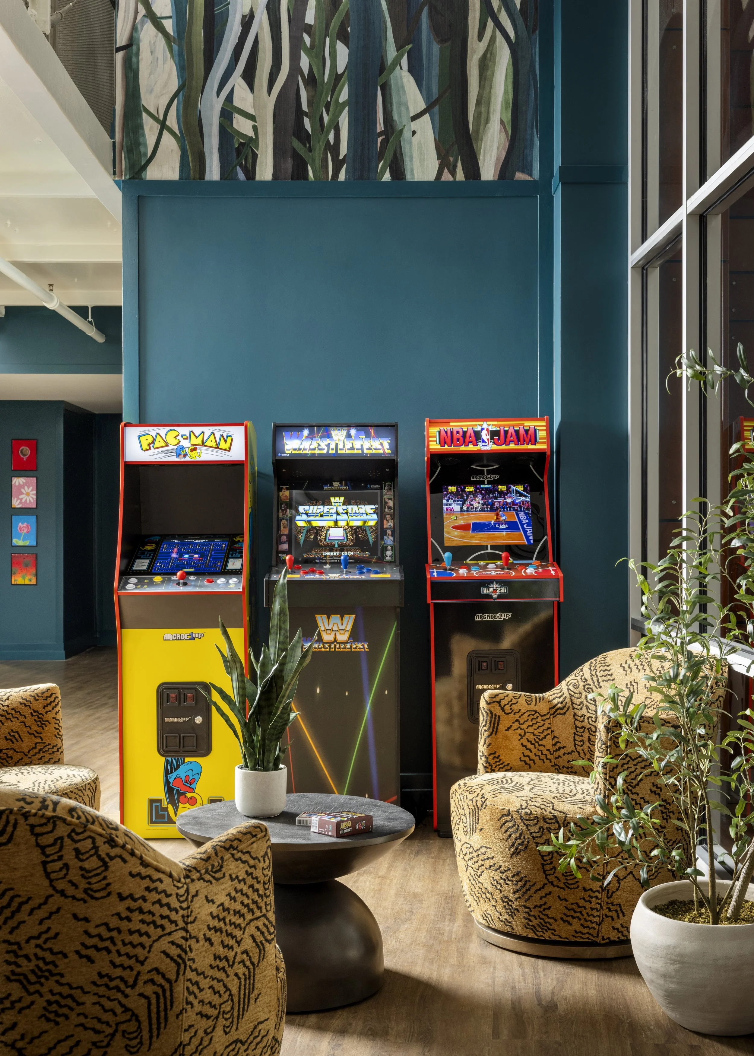 Indoor arcade area with three arcade game machines, a small round table with a potted plant and a deck of cards, surrounded by patterned chairs and green plants. There is a large blue wall and ceiling with partial views of a staircase and windows.