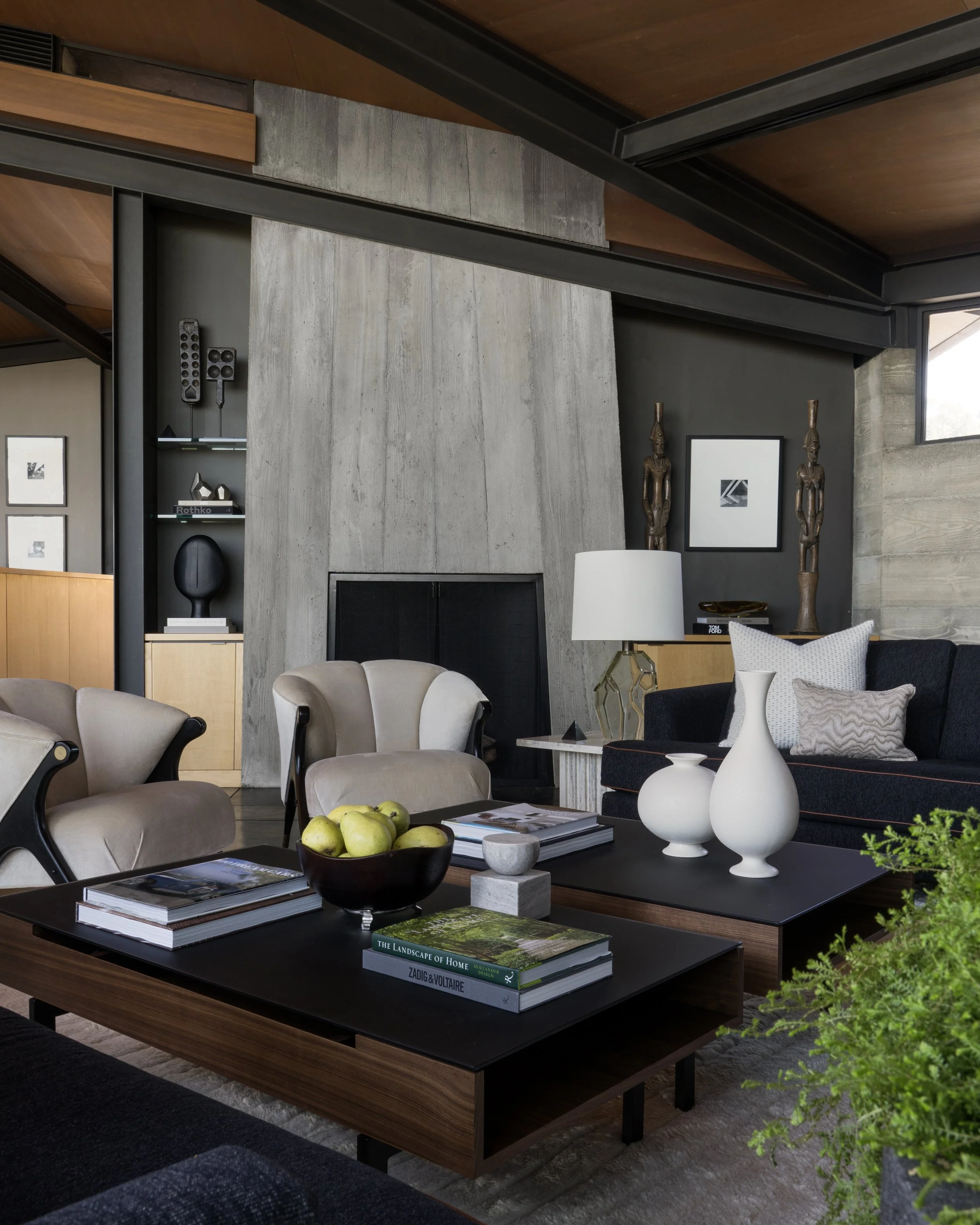 A modern living room with a concrete wall, two tall wooden statues, a black fireplace, and a dark wooden coffee table. The table has a bowl of green apples, several books, and two white decorative vases. There are beige and black seating and cushions
