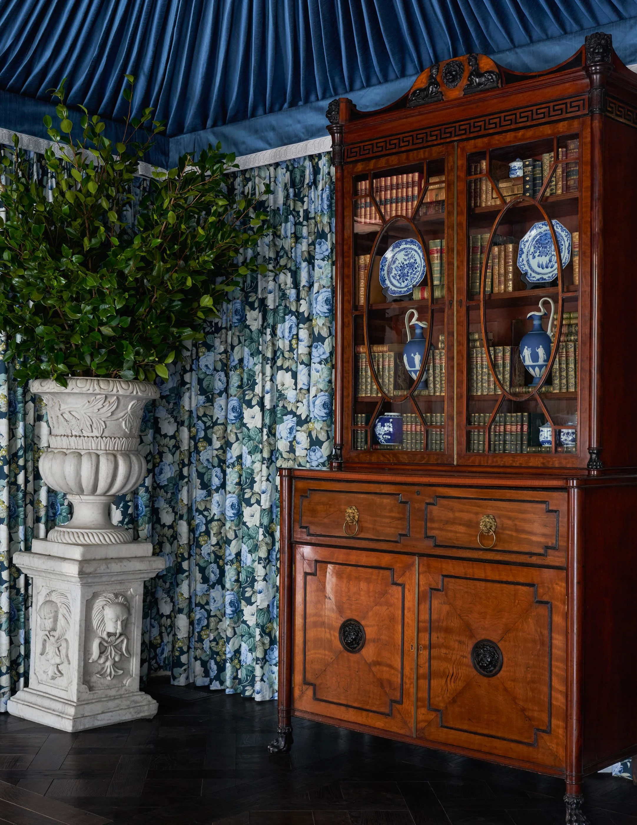 An elegant interior room with a large antique wooden bookshelf filled with old books and blue-and-white porcelain vases, a large white pedestal with a decorative bowl planter, and dark hardwood floors. Blue drapes with floral pattern in the backgroun