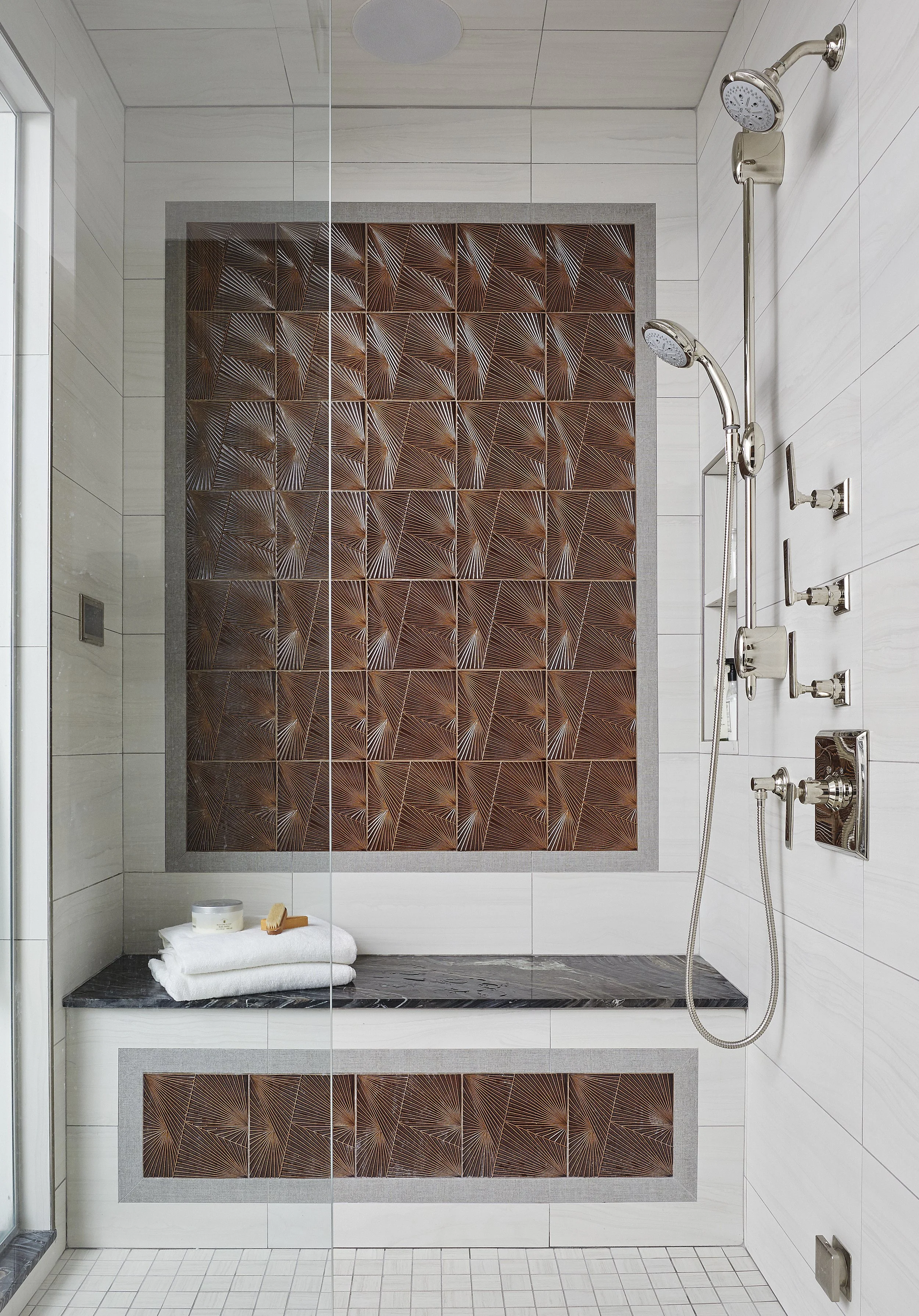 Shower with decorative brown tiles on the wall, white tiled walls, shower fixtures including hand shower and controls, a small shelf with toiletries, and a stack of white towels with a box of soap on top.