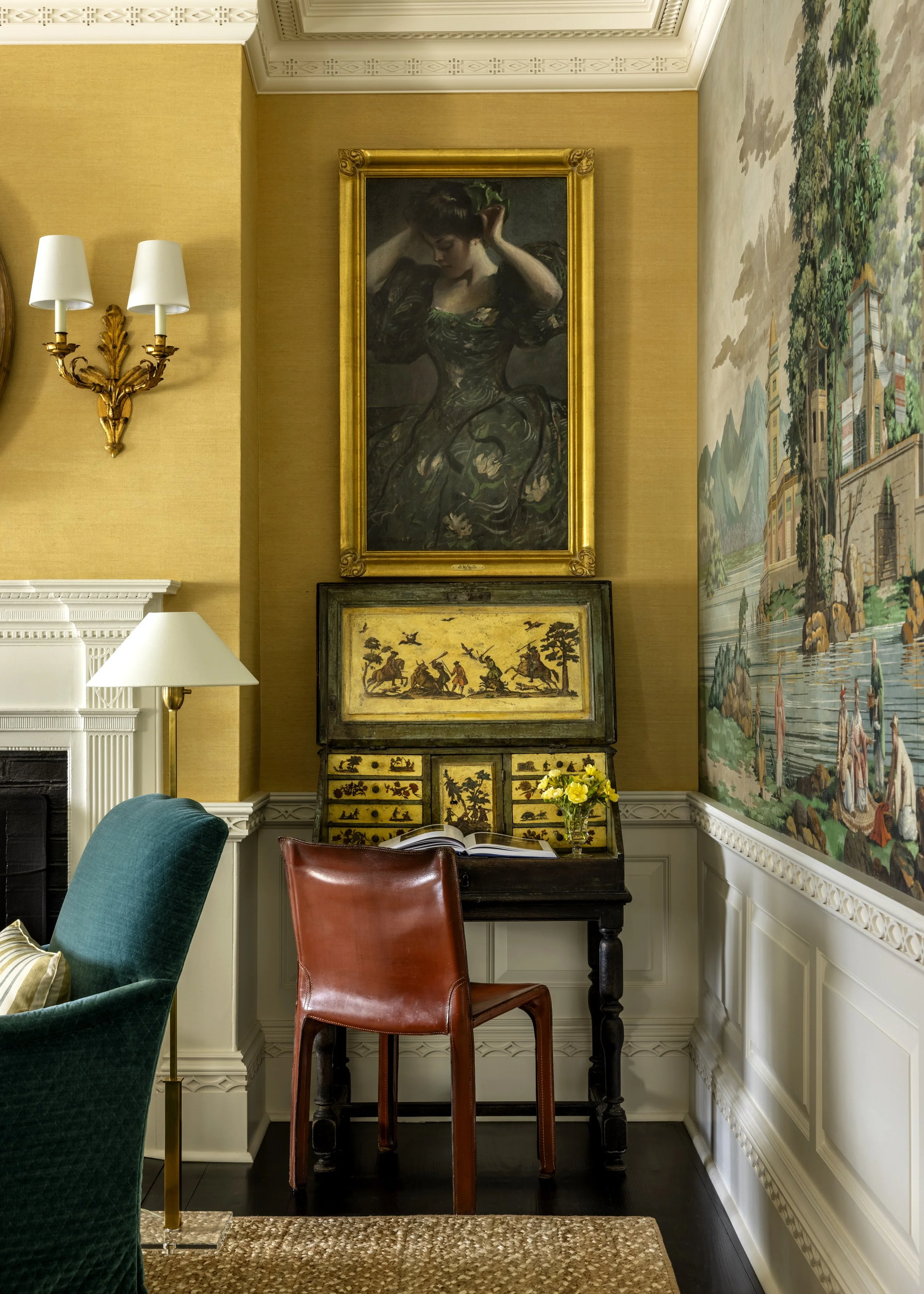 Interior of a room with yellow walls, featuring a large framed portrait of a woman with short brown hair, wearing a dark dress with green and floral details, holding her head with one hand. Below the portrait is an antique cabinet with painted scenes