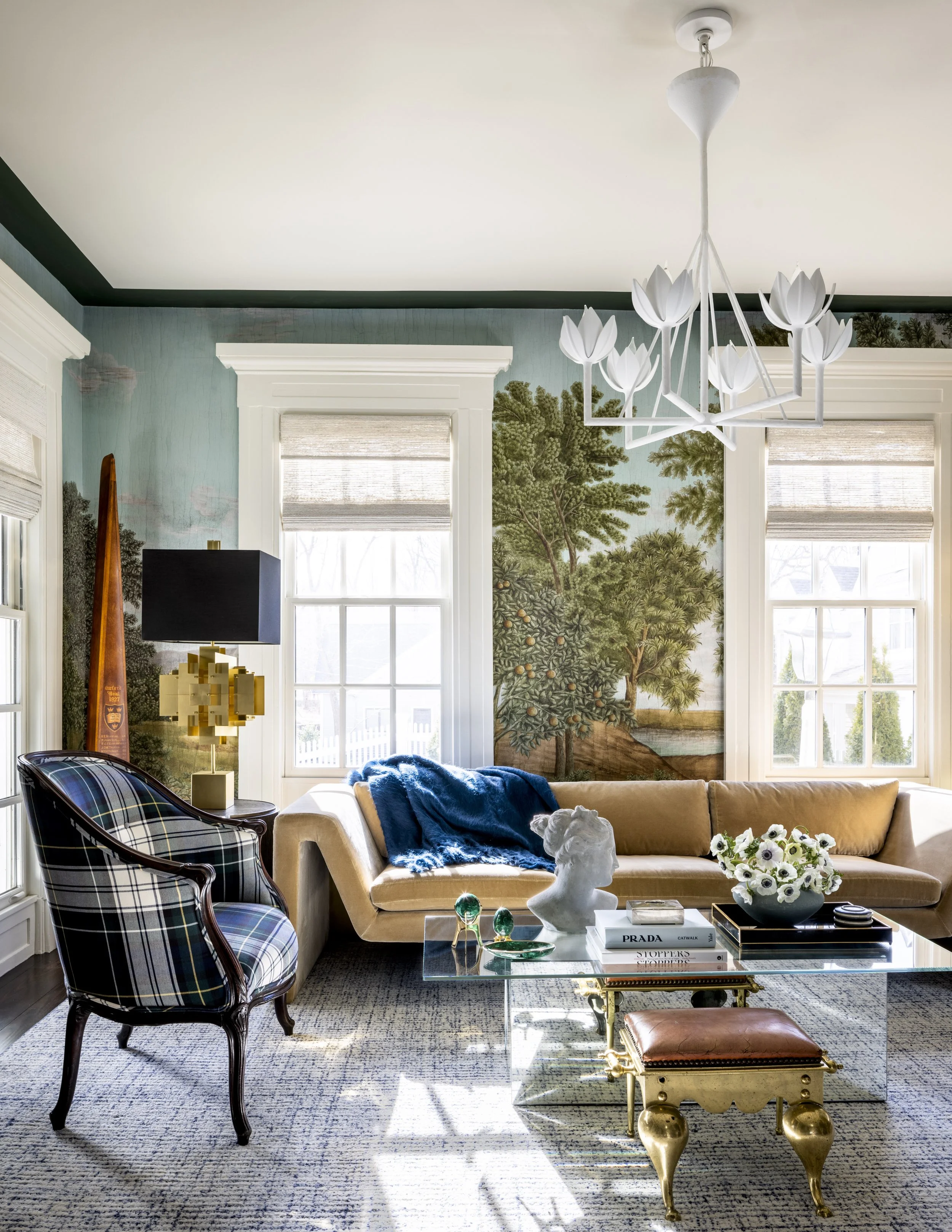 Living room with a beige sofa, a black and white patterned armchair, a glass coffee table with decorative items, and a colorful wallpaper mural of trees on the wall. White windows with blinds, a white chandelier with floral design, and a patterned ru