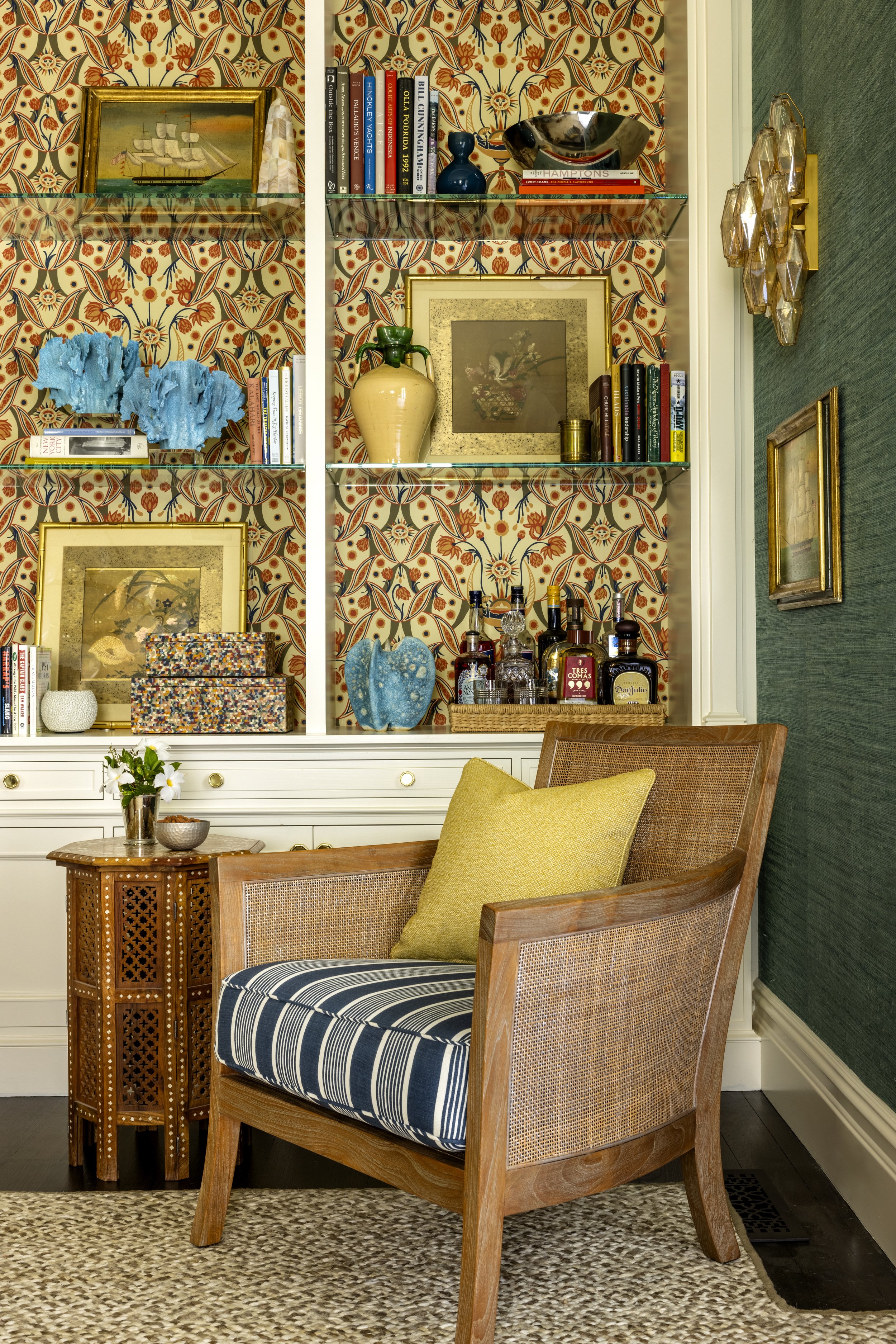 A cozy sitting area with a wooden armchair with a yellow pillow and striped cushion, a small wooden side table with a vase of white flowers, and a beige and brown textured rug. Behind the chair, a white display cabinet with decorative objects, books,