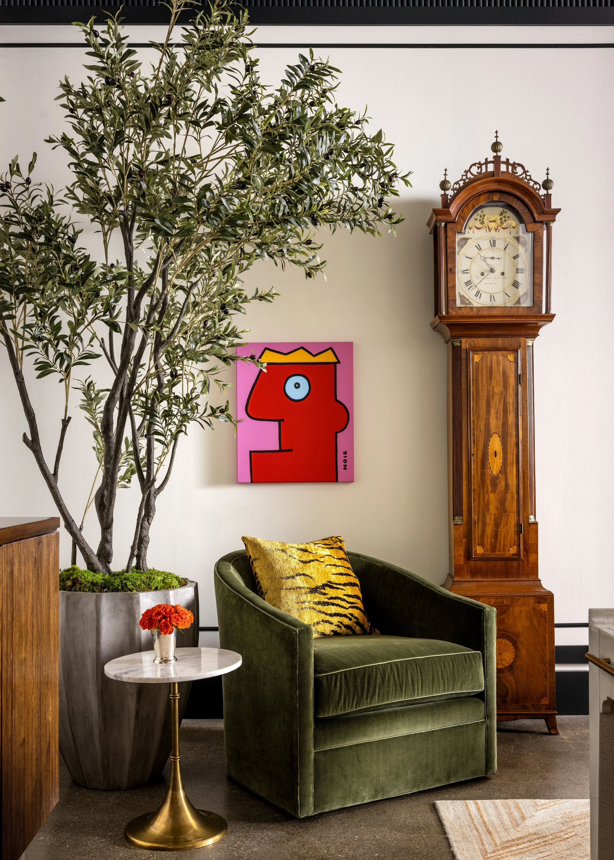 Living room corner with a large potted olive tree, a green velvet armchair with a tiger print pillow, a small white marble side table with a vase of red roses, a vintage wooden grandfather clock, and a pink abstract art piece on the wall.