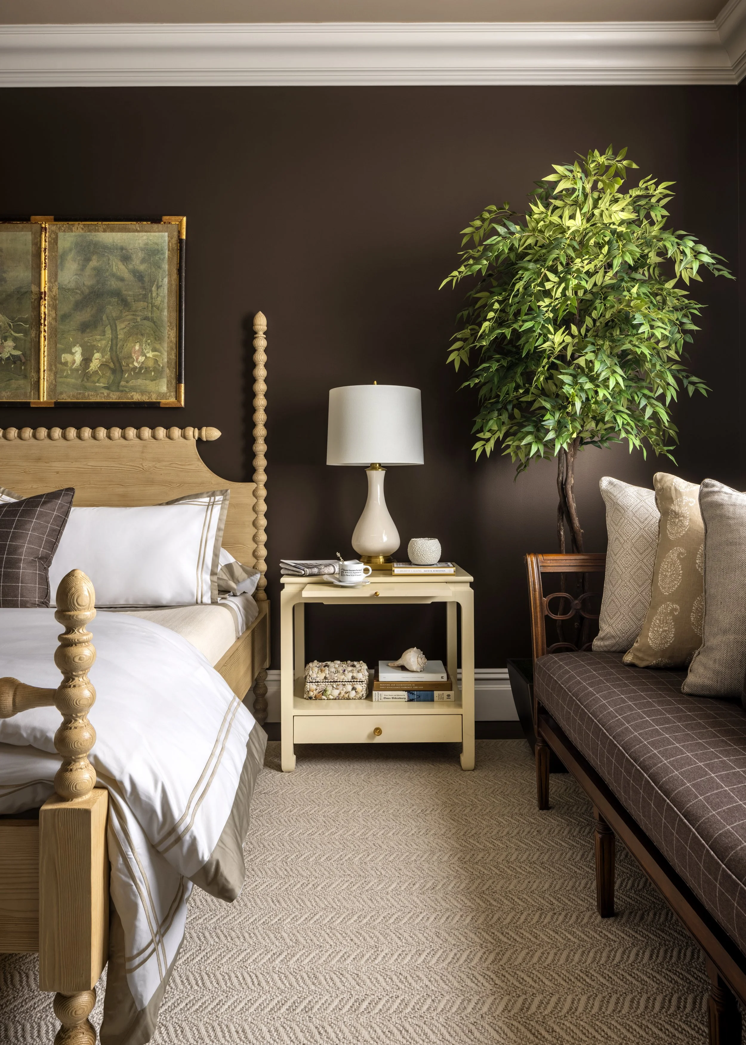 A cozy bedroom corner with a wooden bed frame, white bedding, decorative pillows, a white nightstand with books, a lamp, and a large leafy green indoor plant against a dark wall, with a framed painting hanging above the bed.