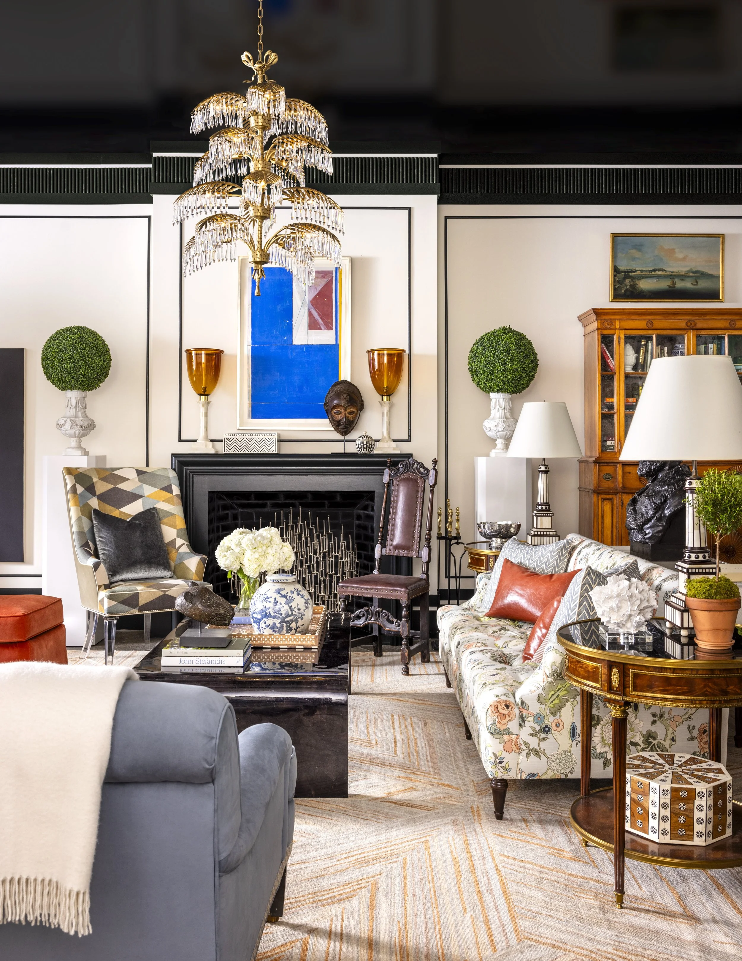 Living room with a chandelier, abstract art, green topiary plants, fireplace, various chairs and sofas, and decorative items.