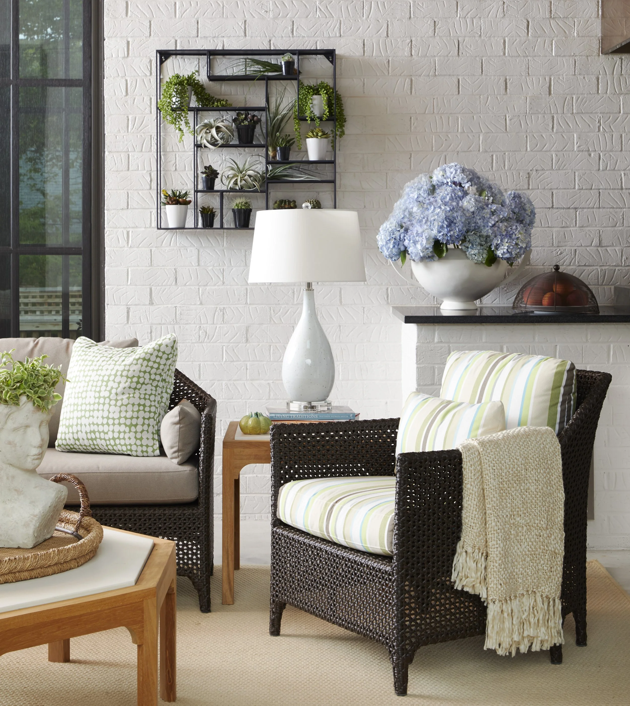 Living room corner with a white brick wall, a black metal wall shelf holding various potted green plants, a white ceramic planter on a small table with a large bouquet of blue hydrangeas, a white table lamp, a wicker basket with apples, a beige woven