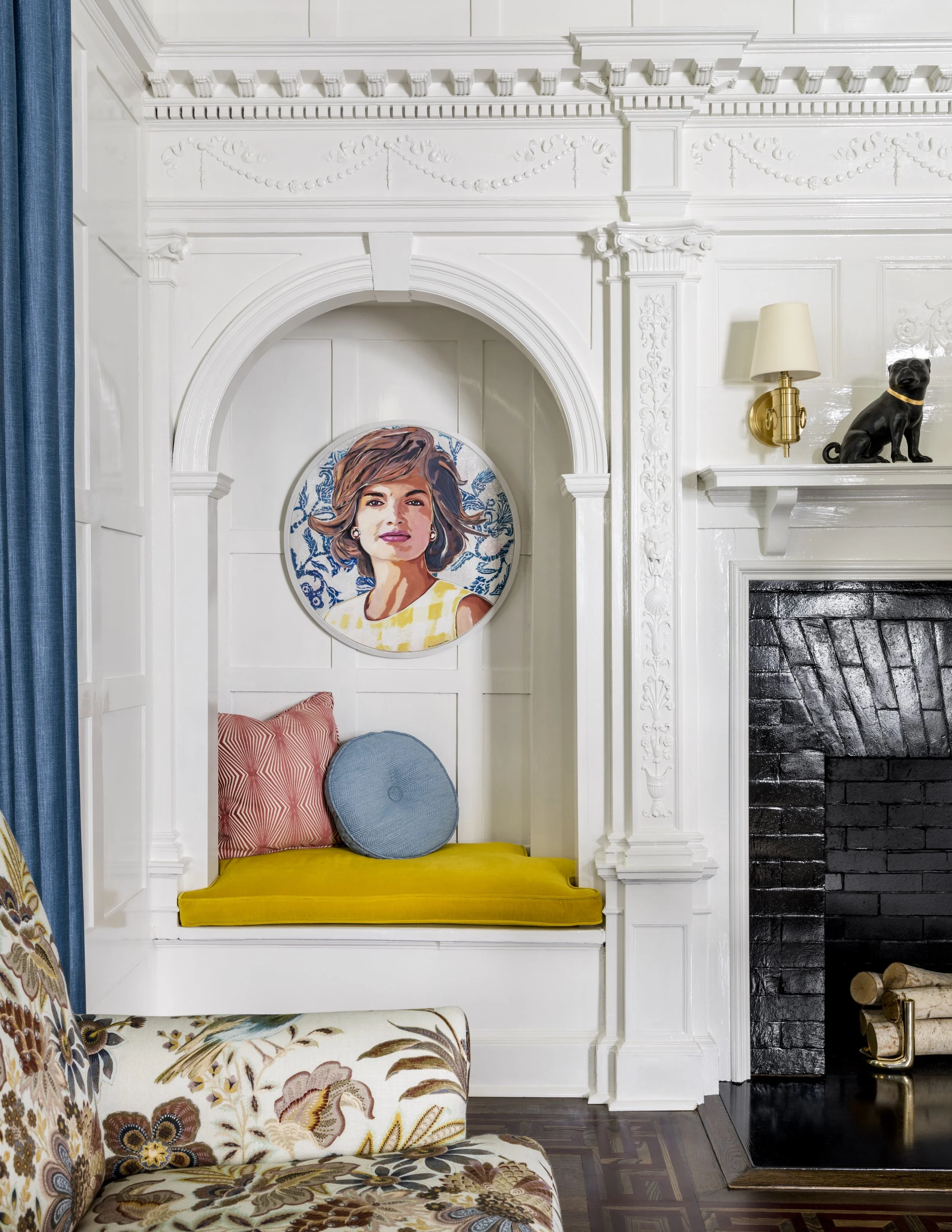A white ornate fireplace with decorative moldings and a mantel. Inside the fireplace is a yellow bench with two round pillows, one blue and one patterned, and a portrait of a woman on the wall behind it. To the right of the fireplace is a black brick