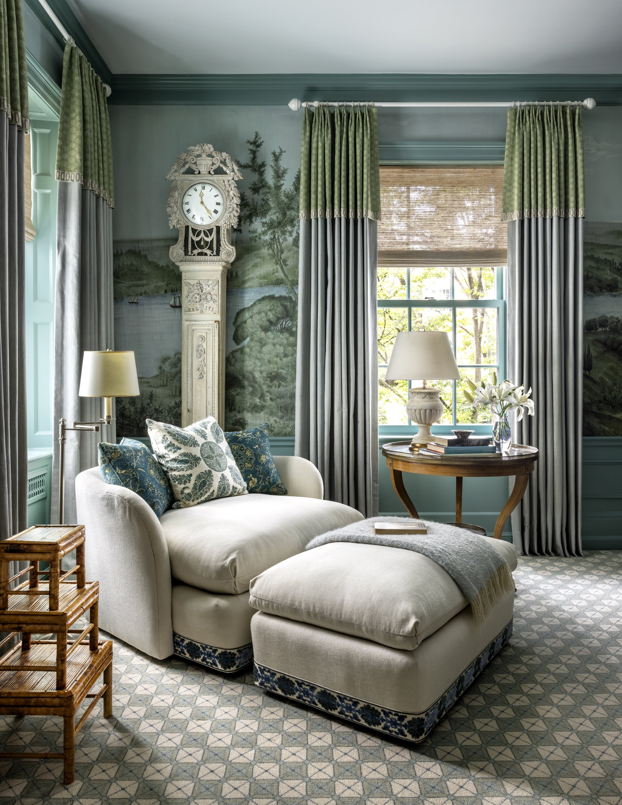 Living room with large window, green curtains, a white sofa with blue and white patterned pillows, a wooden side table, a table with books and a lamp, a vintage clock, and a mural mural depicting a landscape with trees and water.