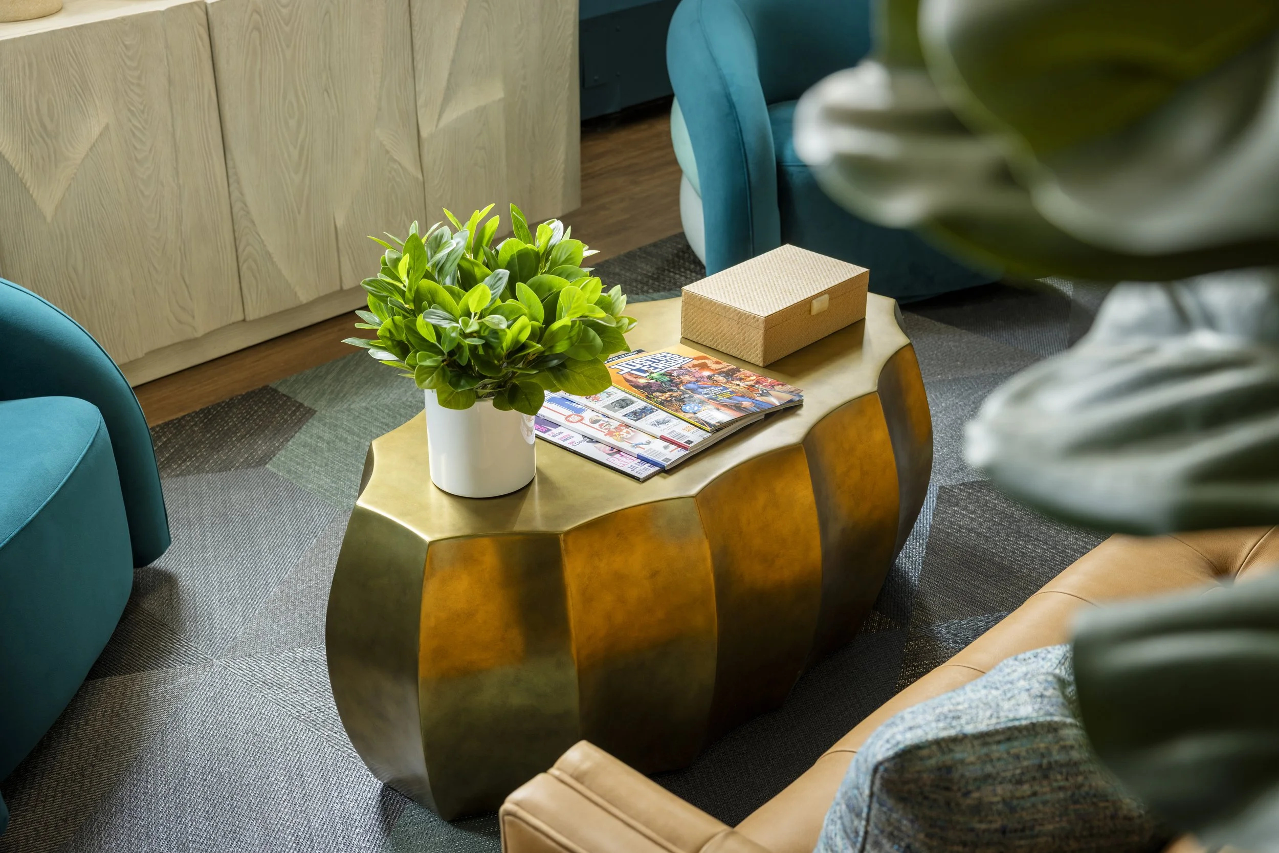 A round metallic coffee table with a green potted plant, magazines, and a box on top, in a cozy room with a wooden cabinet and teal upholstery.