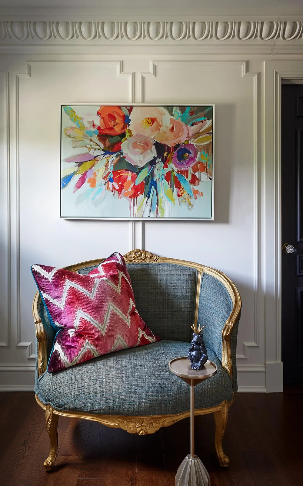 An interior living space with a vintage style sofa featuring gold trim, a pink and white chevron pillow, a small side table with a decorative figurine, and a colorful floral painting on a white paneled wall.