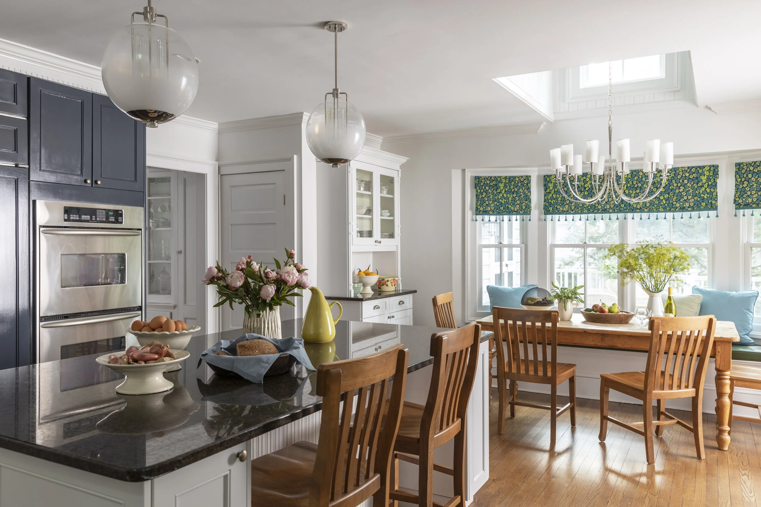 A bright kitchen with white walls and large windows with floral valances, a dark countertop island with wooden chairs, a floral arrangement, and a dining area with a chandelier.
