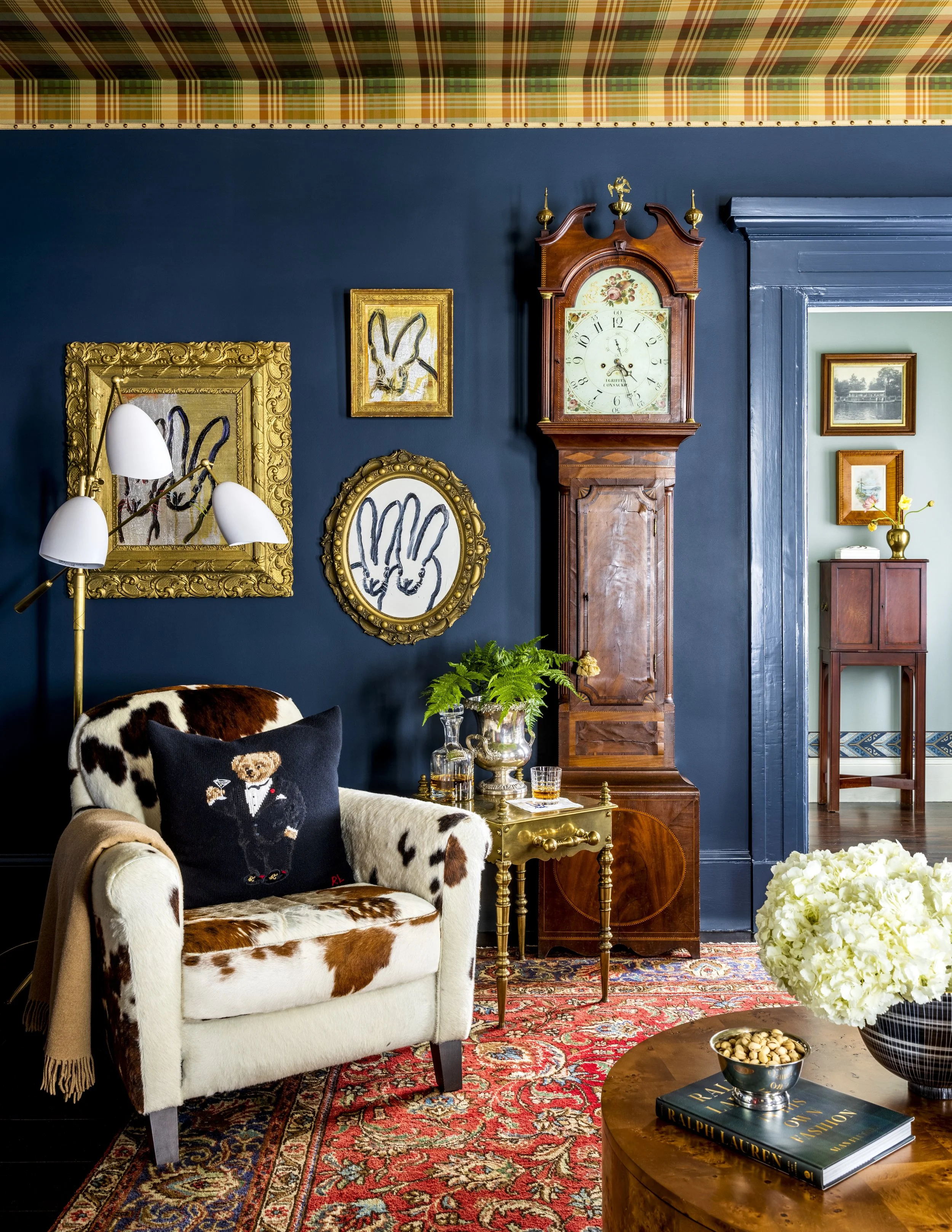 A cozy living room corner with a white armchair covered with cowhide fabric, a black pillow with a dog embroidered in a tuxedo, and a beige shawl draped over the armrest. A gold-framed painting and a round mirror with a gold frame hang on a deep blue