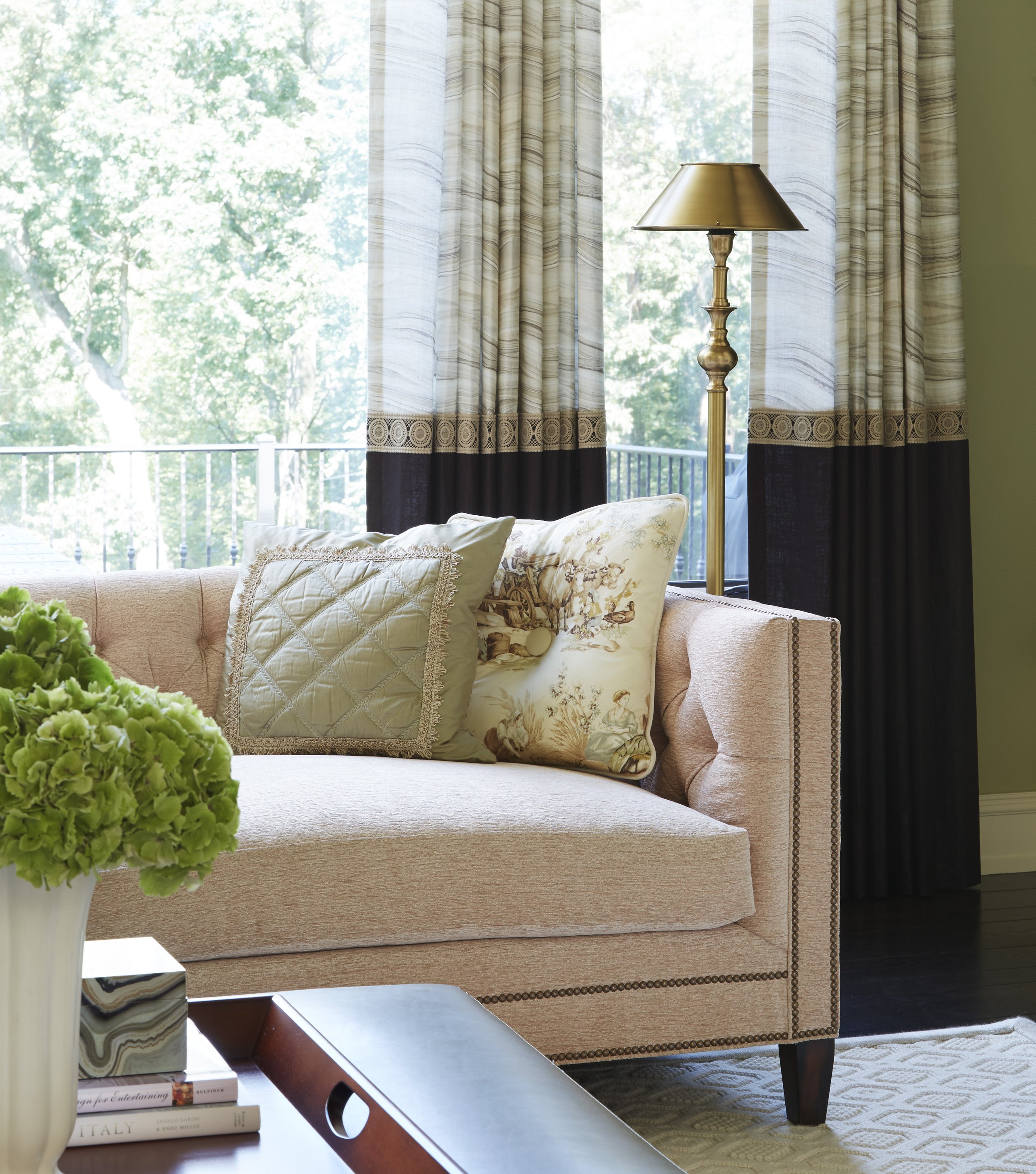 Living room with beige upholstered sofa, decorative pillows, a wooden side table with books, a floor lamp with a brass finish, large windows with beige and black curtains, and a vase of green hydrangeas in the foreground.