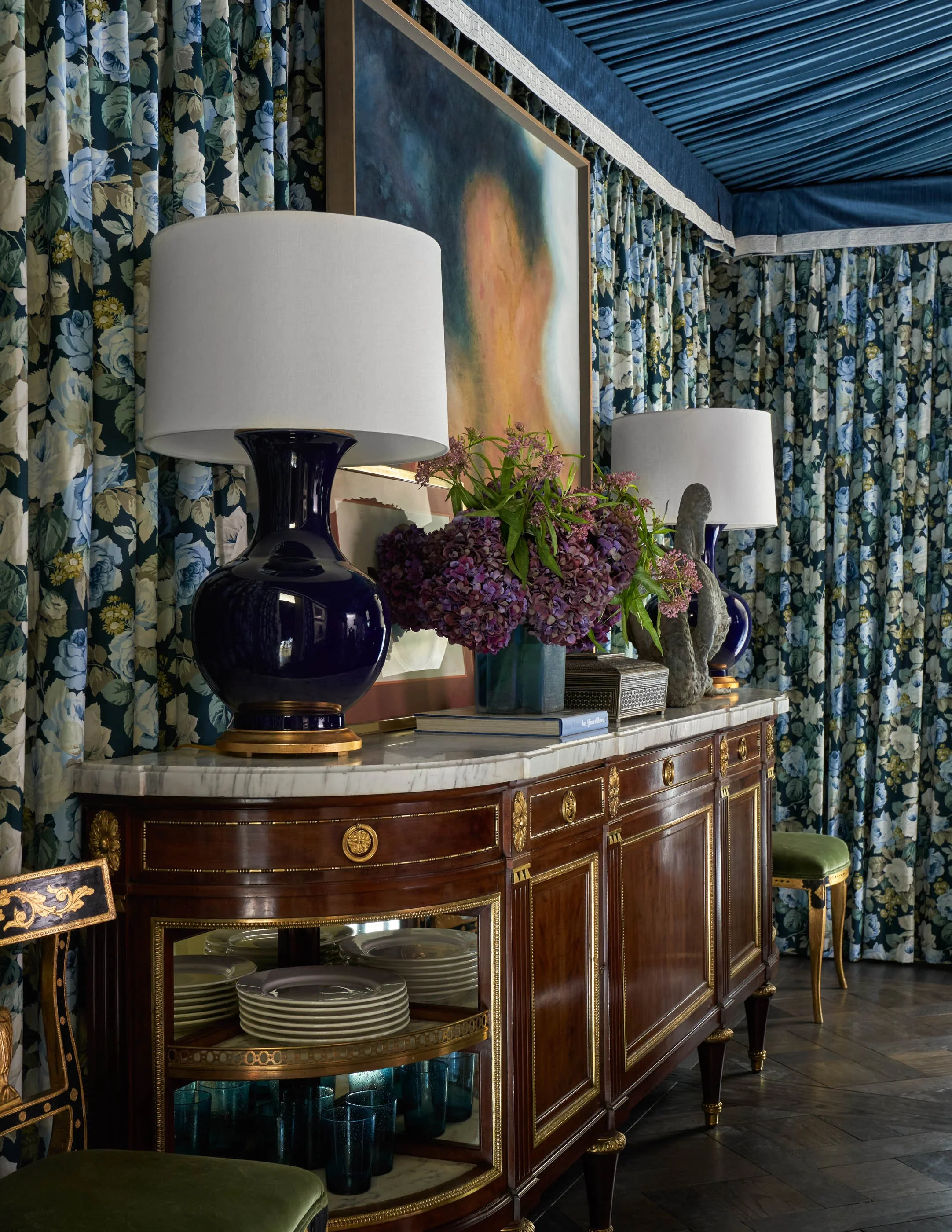 A vintage wooden sideboard with gold accents and a marble top, featuring a glass-front display with plates inside. On top of the sideboard are two black lamps with white lampshades and a floral arrangement of purple and green flowers. The background 