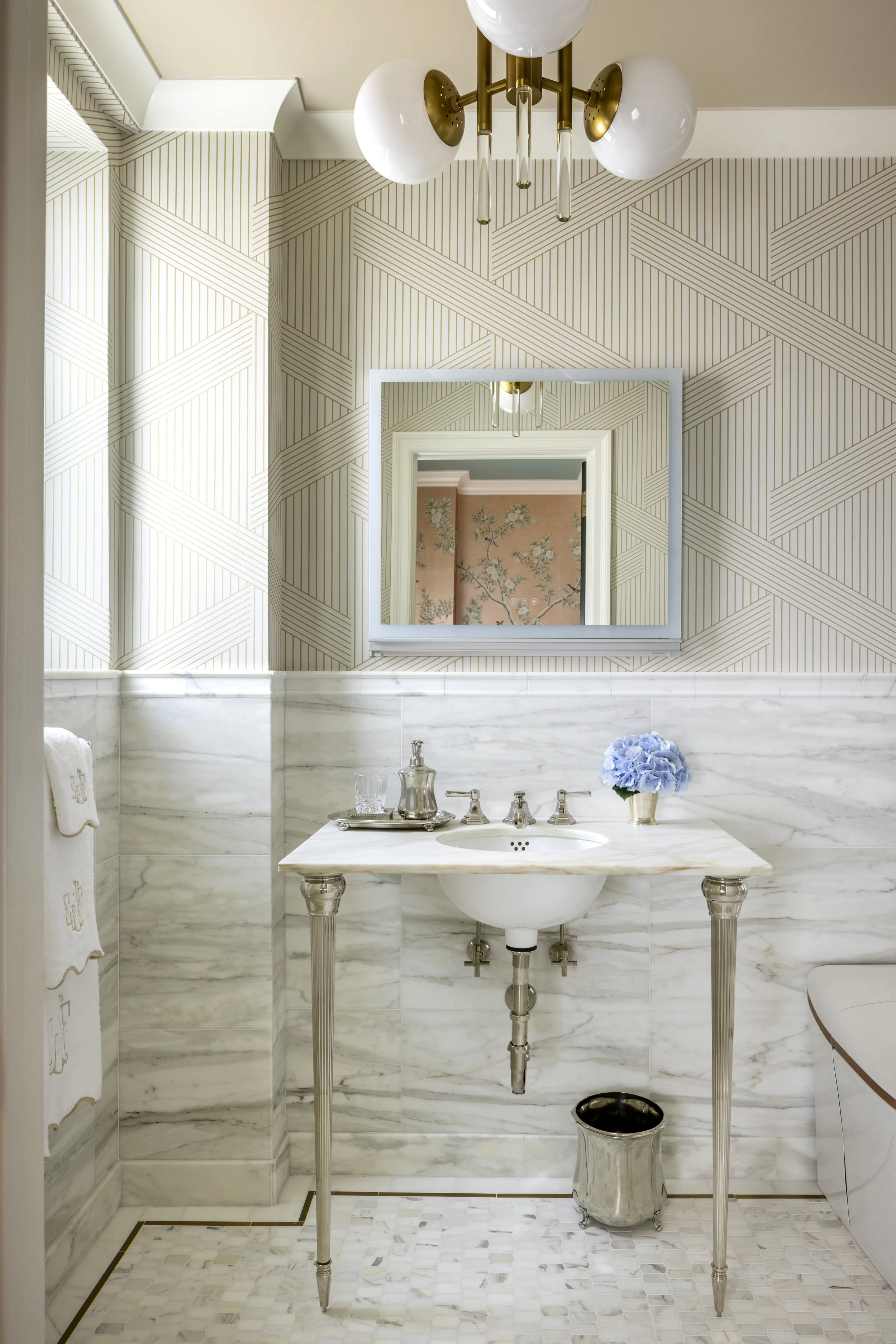 Bathroom with marble walls and floor, featuring a marble sink with decorative silver legs, a mirror, a chandelier, a bouquet of blue hydrangeas, and tiled decor.