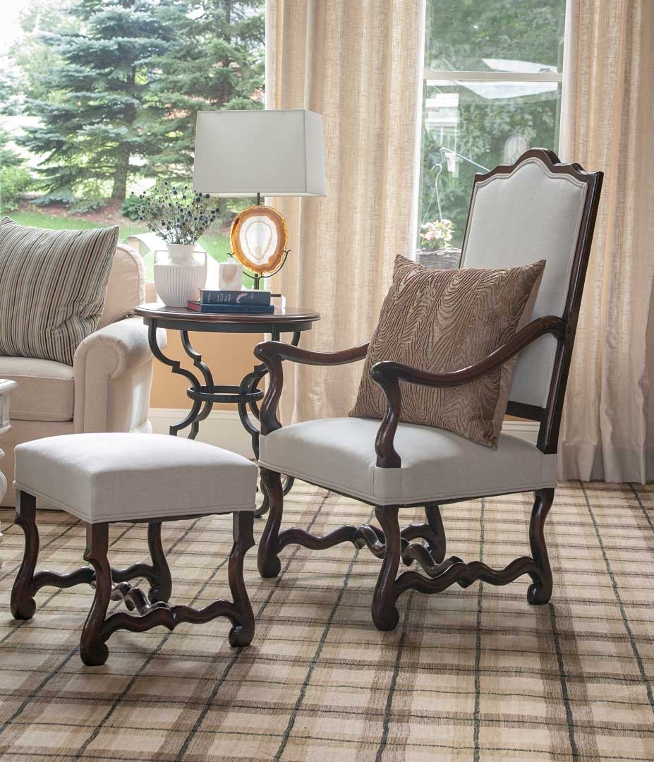 Living room corner with white upholstered armchair with wooden frame and a matching ottoman, a side table with a table lamp, books, and a decorative object, and a beige sofa with striped pillows, large window with curtains, and a view of green trees 