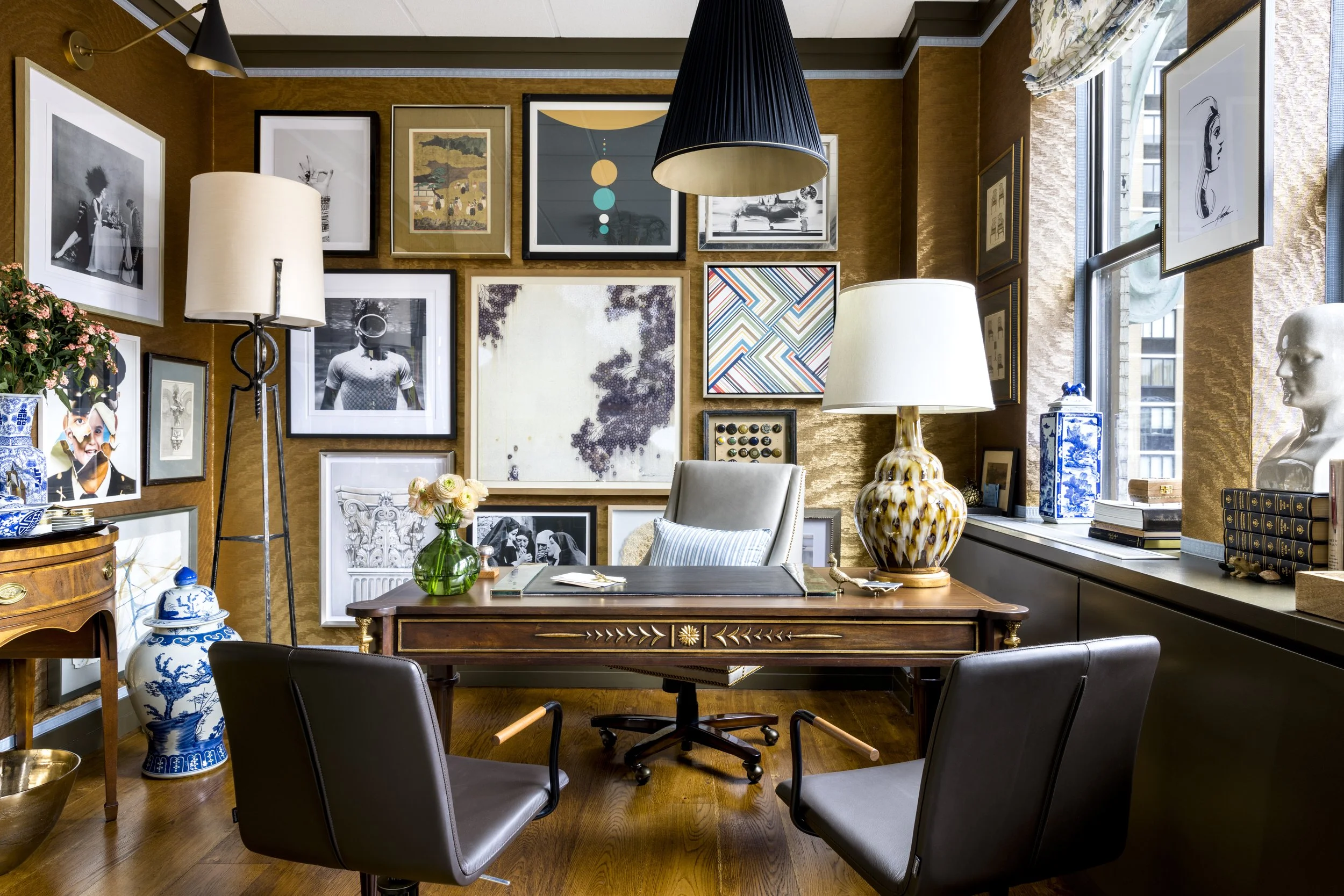 A cozy, art-filled dining room with a wooden table, black chairs, a variety of framed artwork on golden-brown walls, and decorative lamps. There are flower arrangements and vases, and a large window with patterned curtains letting in natural light.