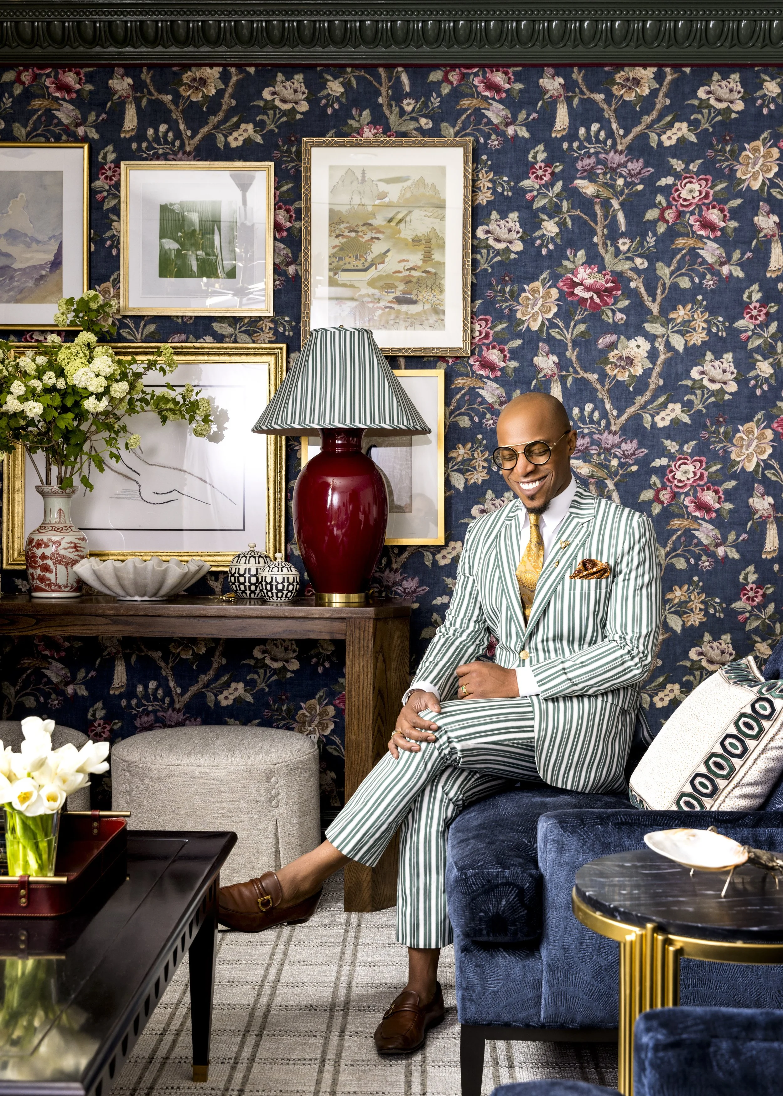Corey Damen Jenkins dressed in a green and white striped suit, sitting on a blue velvet sofa, smiling with glasses, in a decor-filled room with floral wallpaper, artwork, a red vase lamp, and a table with flowers and decorative items.
