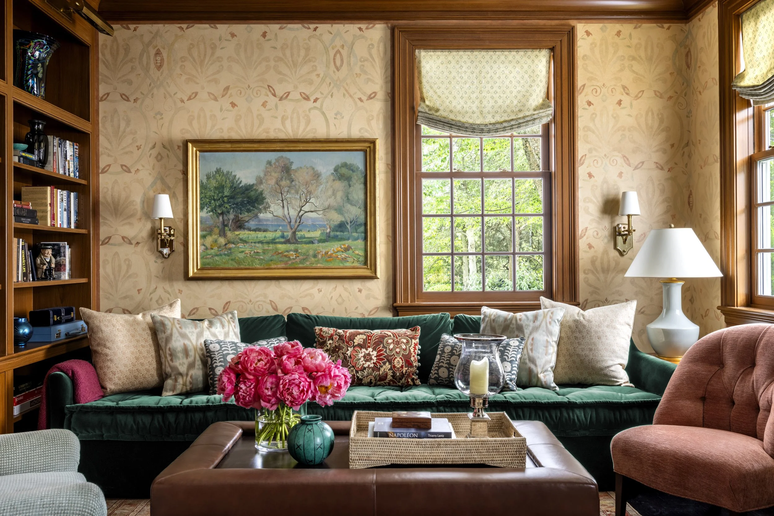 Living room with a bookshelf on the left, a painting of trees above a green velvet sofa, and a window with a cream-colored blind. The room has beige wallpaper and wooden trim, with various lamps and decorative pillows, and a vase of pink flowers on t