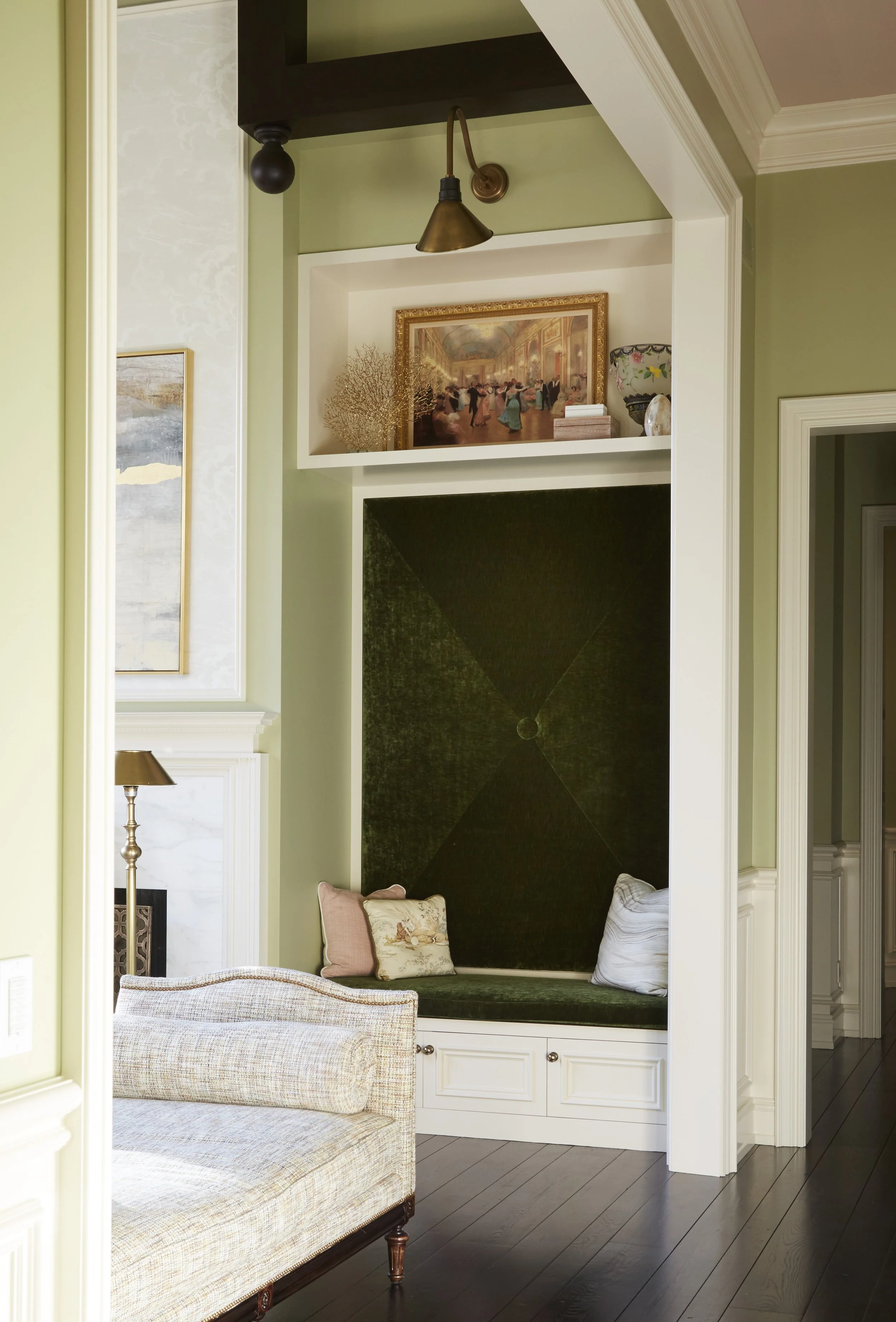 A living room corner with a built-in cushioned window seat with pillows, dark hardwood floor, and white cabinetry underneath, adjacent to a white wall with framed artwork and a table lamp.