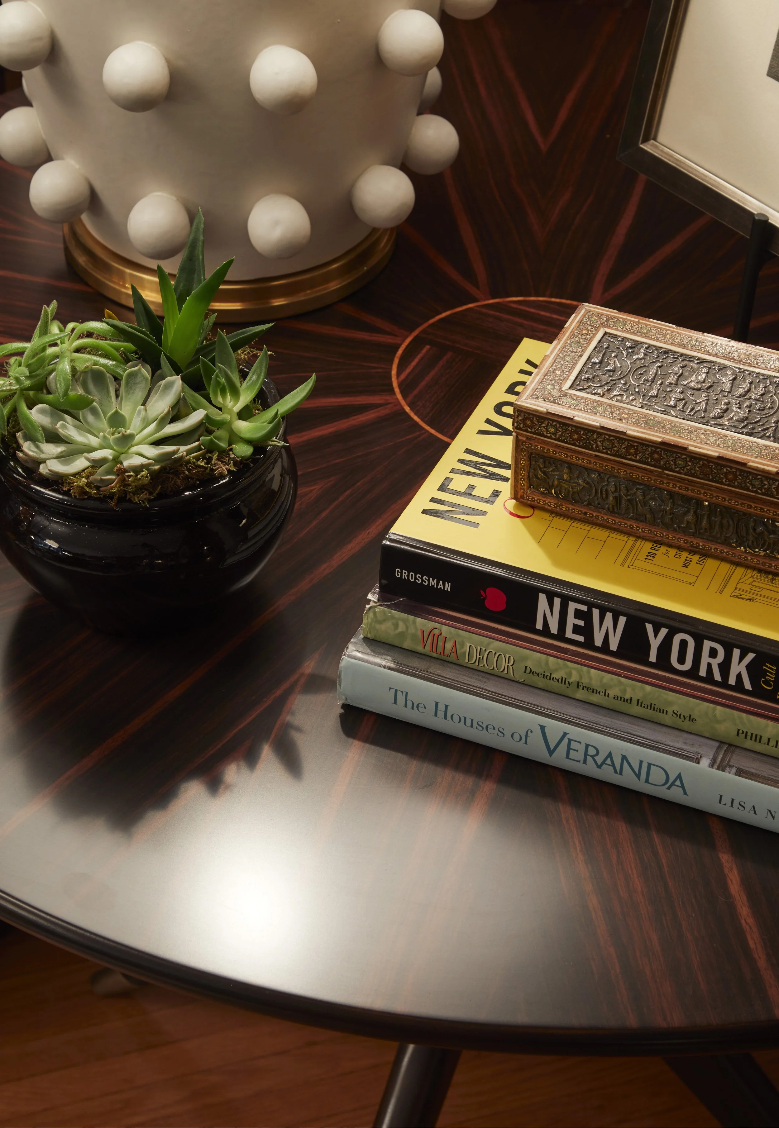 A close-up of a dark wooden table with a potted succulent plant, a decorative box with intricate metalwork, and a stack of books about New York City and Verona. In the background, part of a white decorative lamp and a framed picture are visible.