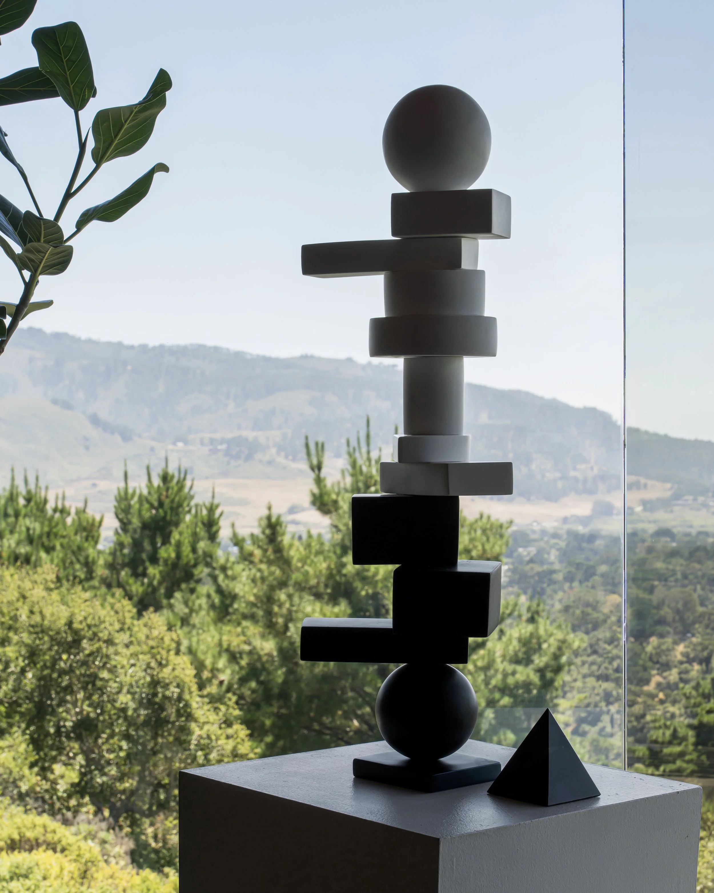 Abstract sculpture consisting of stacked geometric shapes including spheres, cubes, and rectangular blocks, placed on a pedestal with a window view of mountains and trees in the background.