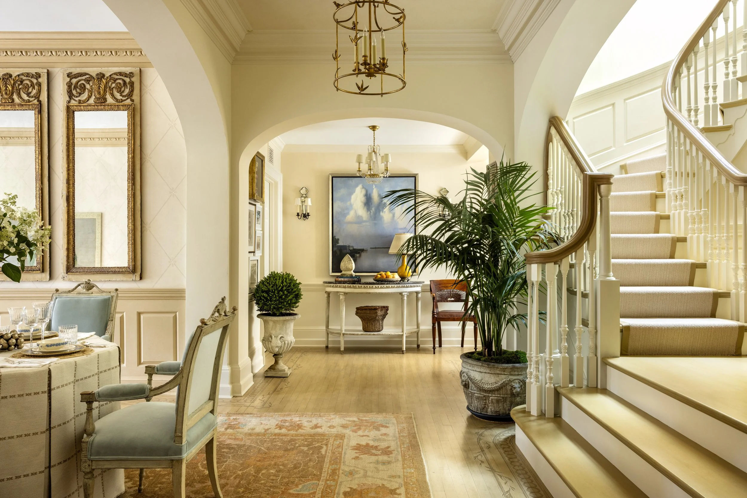 Elegant interior of a home with a staircase, large plants, framed artwork, and chandelier lighting.
