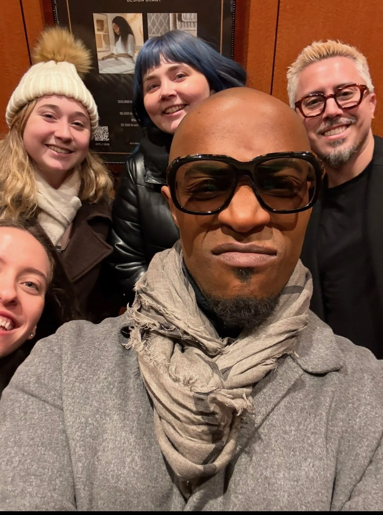 Group of six people posing for a selfie indoors. The person in front is bald, wearing glasses, a light gray hoodie, and a beige scarf. To his left, a woman with light brown hair, wearing a white knit hat with a pom-pom, and a dark coat. Behind him, a woman with short blue hair, smiling, wearing a black leather jacket. To her right, a man with blonde hair, glasses, and a black shirt. Partially visible at the bottom left is another woman with dark hair, smiling. The background features a wooden wall and a framed poster with a picture of a woman reading.
